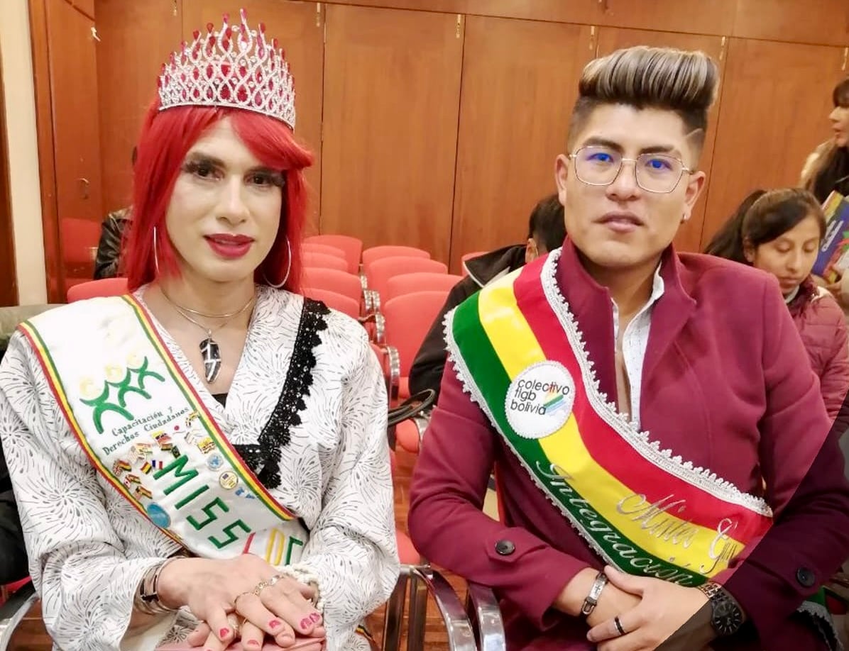 Två personer sitter bredvid varandra i en sal iklädda ordensband över axlarna. Personen till vänster bär en stor glittrande krona och röd peruk, medan personen till höger har glasögon och en vinröd kavaj.
