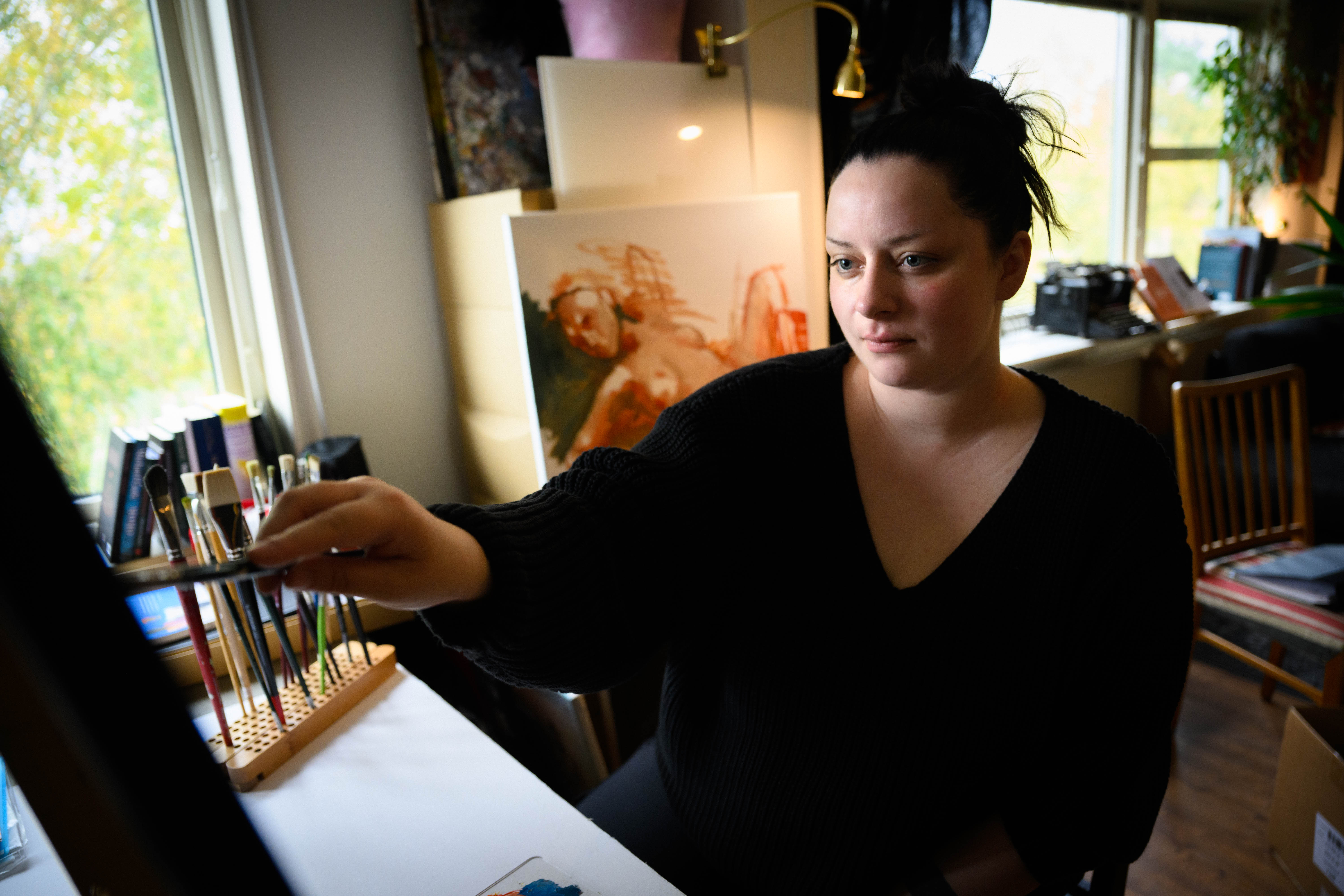 En person i svart tröja målar fokuserat på en duk i en ljus ateljé.
