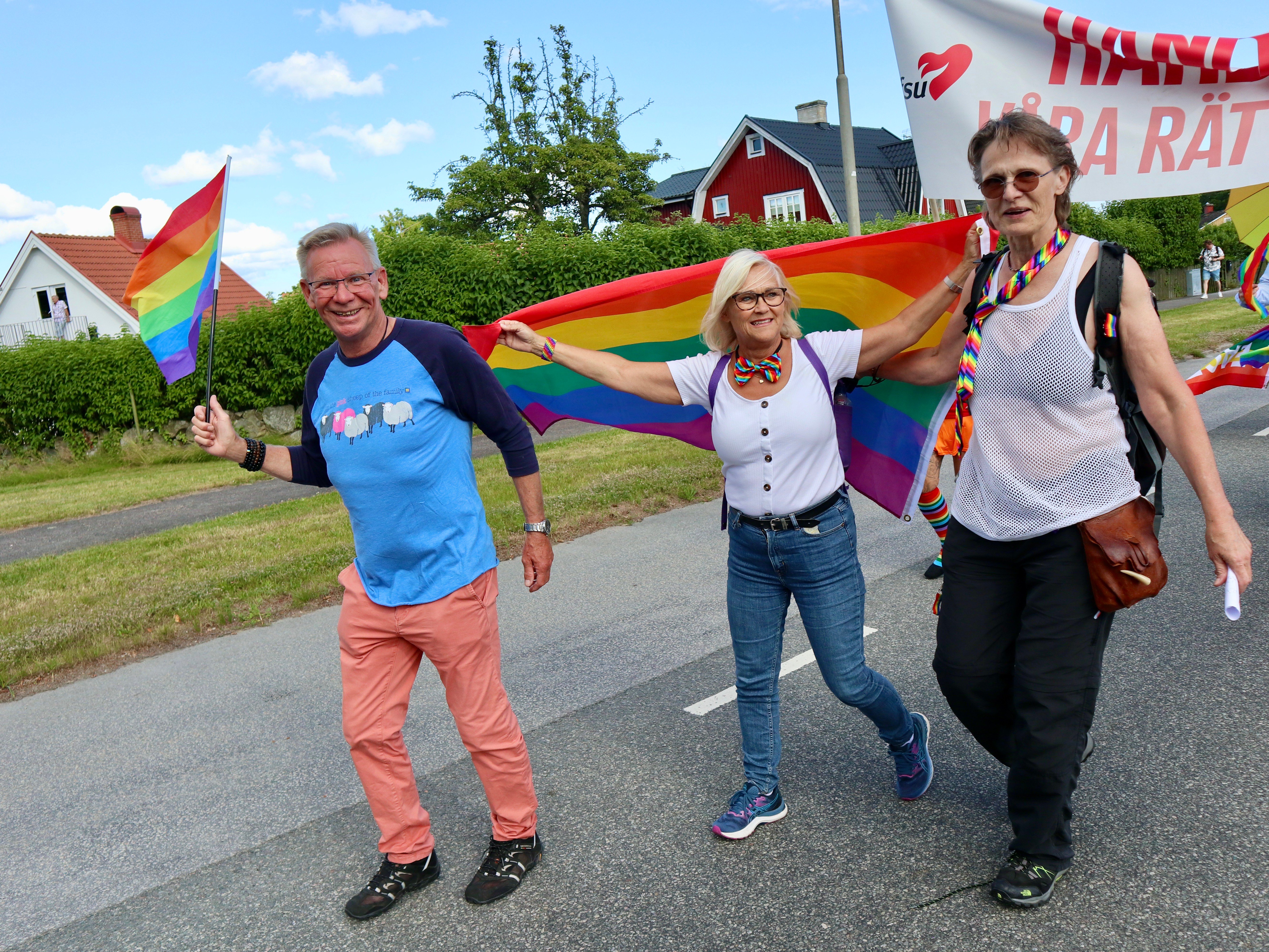 Tre personer går längs en väg i en parad och håller gemensamt upp en stor regnbågsflagga.