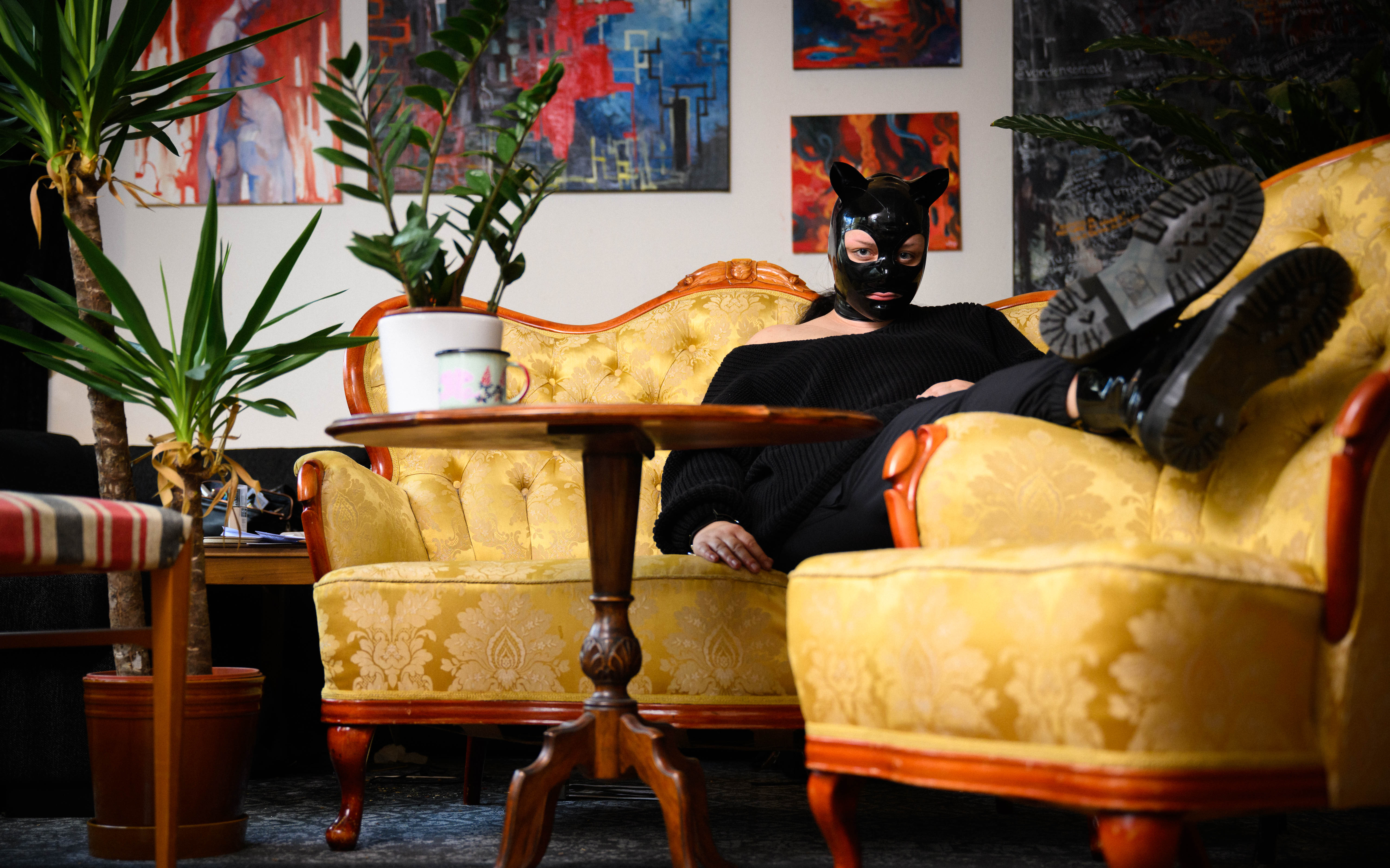 En person i svart mask med öron vilar i en gulmönstrad soffa omgiven av växter och tavlor.