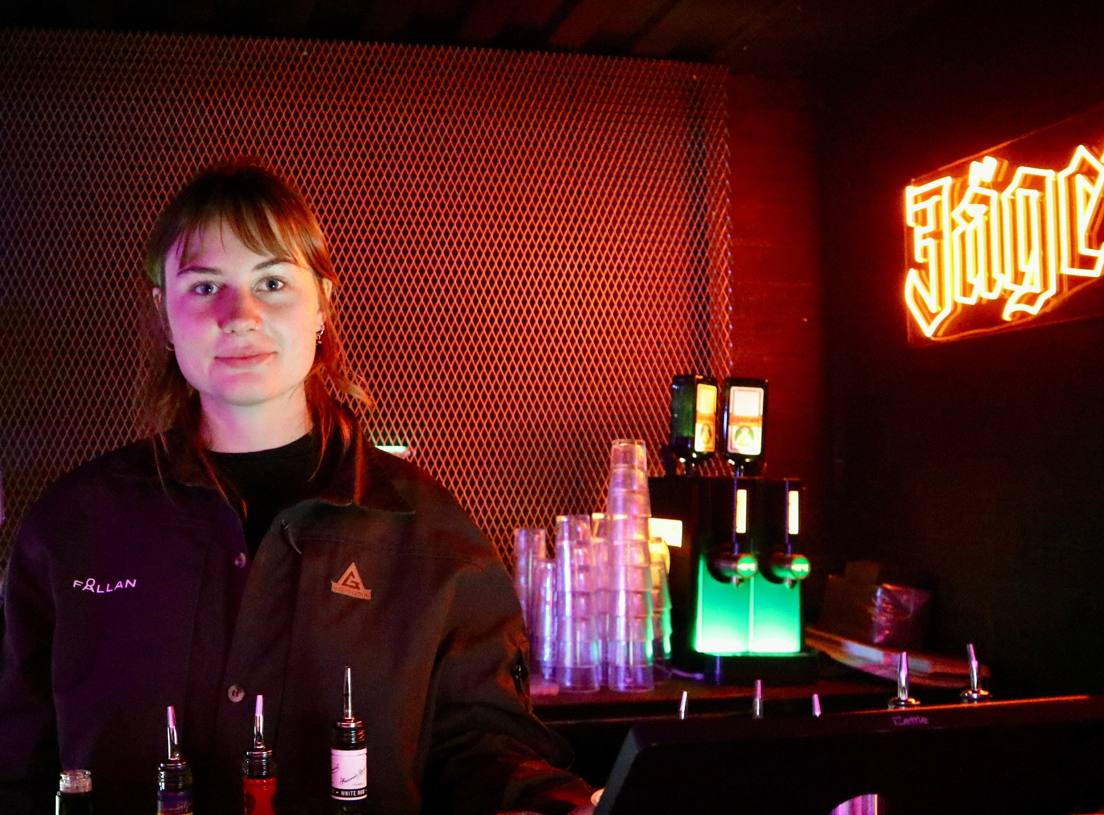 Person i mörk jacka står i en bar med neonljus och flaskor i förgrunden.