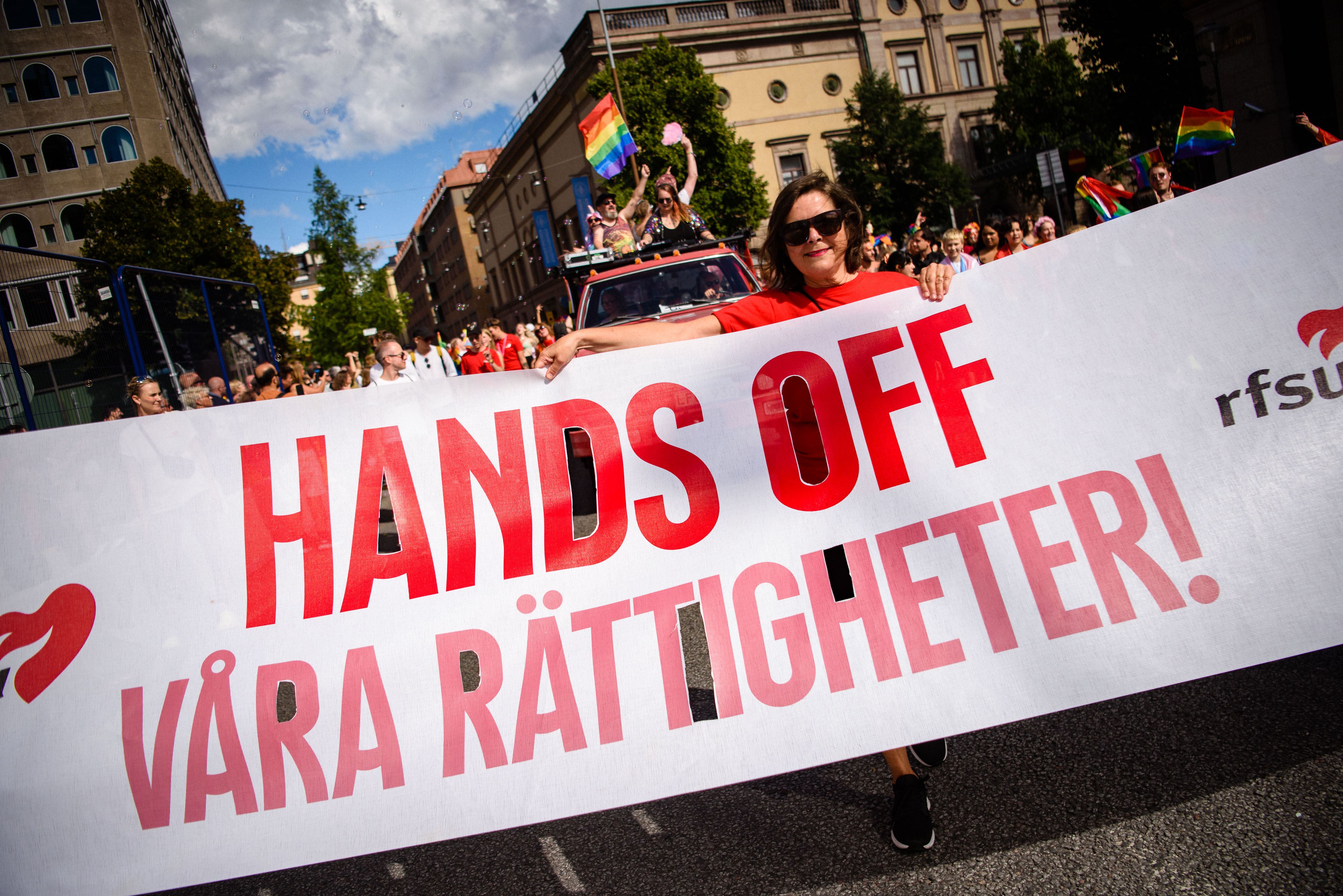 En kvinna i röda kläder och solglasögon går i en Pride-parad och håller i en stor vit banderoll. På banderollen står texten ”HANDS OFF VÅRA RÄTTIGHETER!” i stora röda bokstäver, tillsammans med RFSU:s logotyp. Bakom henne syns en röd pickup med paraddeltagare, regnbågsflaggor och en folkmassa som rör sig längs en stadsgata under en blå himmel med lätta moln.
