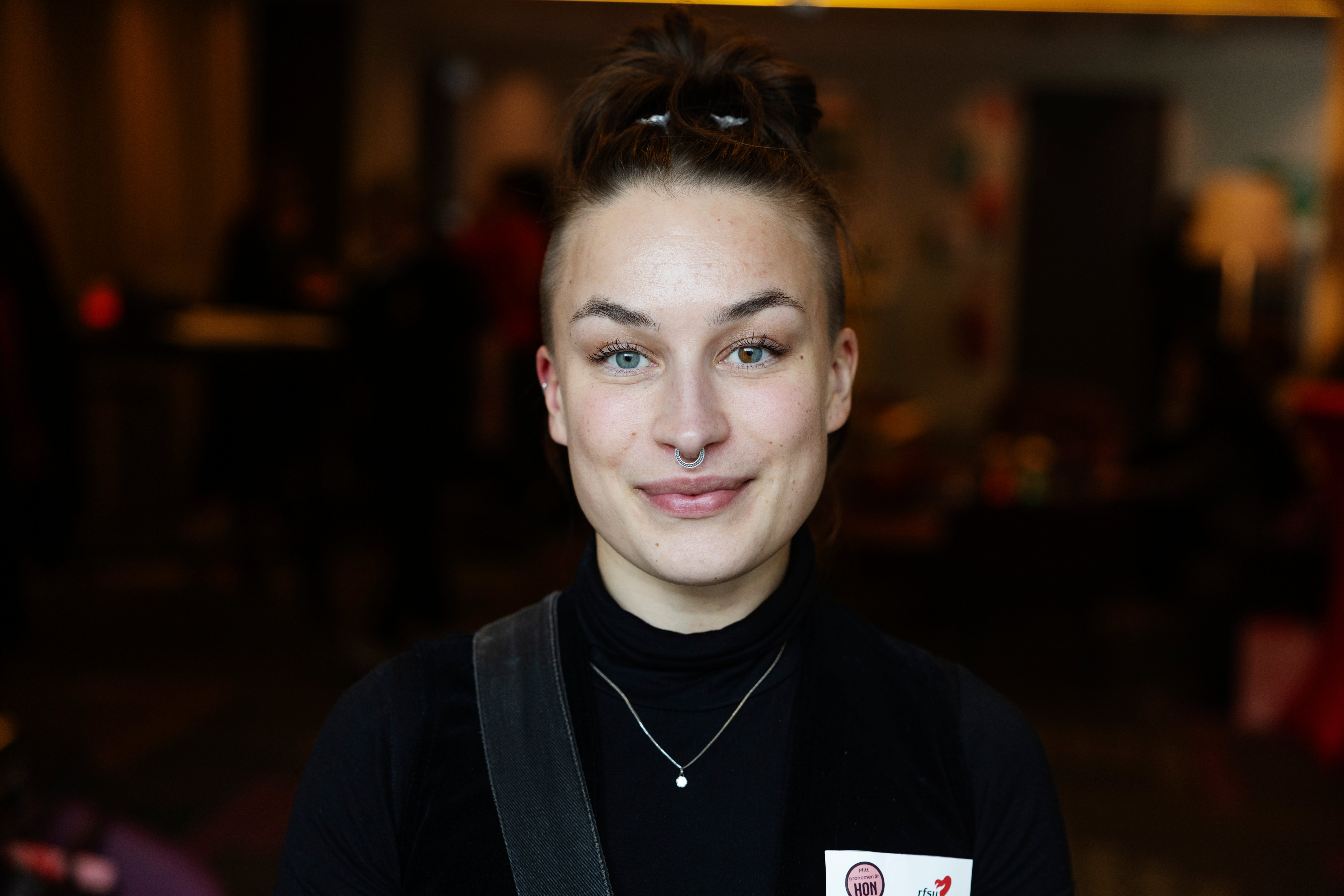 Ett närbildsporträtt på en ung person med uppsatt hår och en näspiercing som blickar lugnt in i kameran.