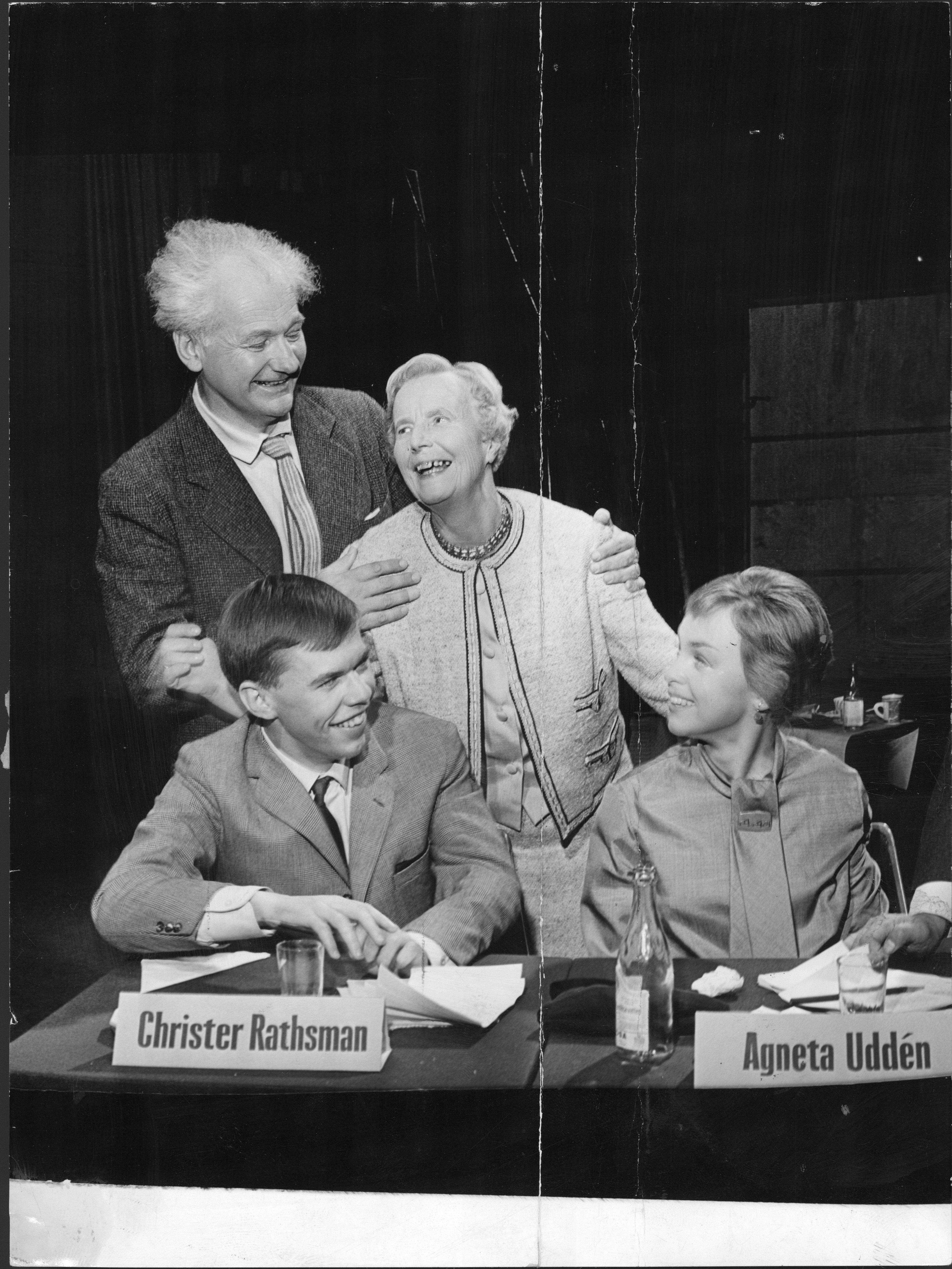 Svartvit bild med en kvinna och en man som står och tittar glatt på varandra. Under dem sitter två ungdomar med namnskyltar där det står Crhister Rathsman och Agneta Uddén på. De ser också glada ut. 