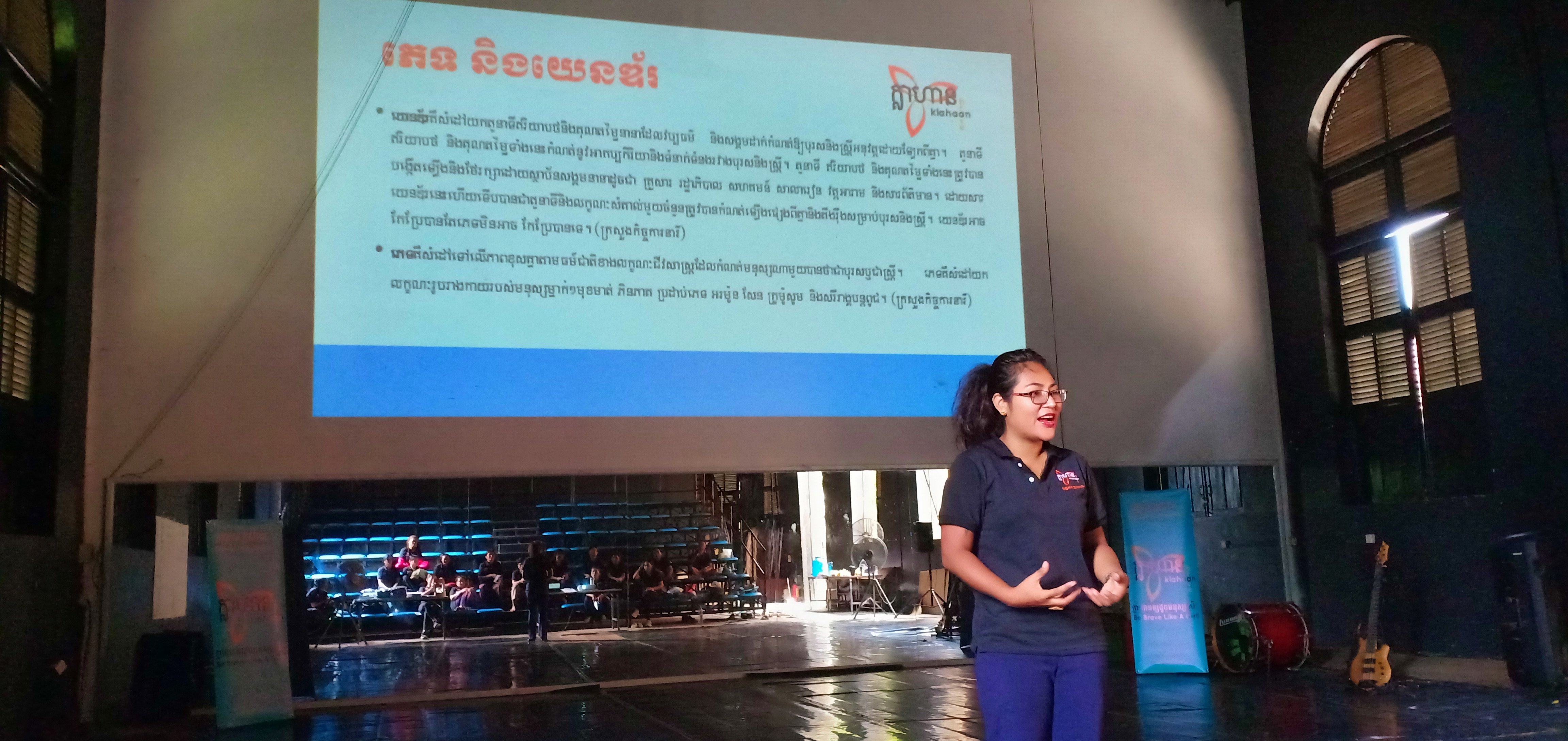En kvinna håller en presentation på en scen framför en stor projektorduk med text på khmer.