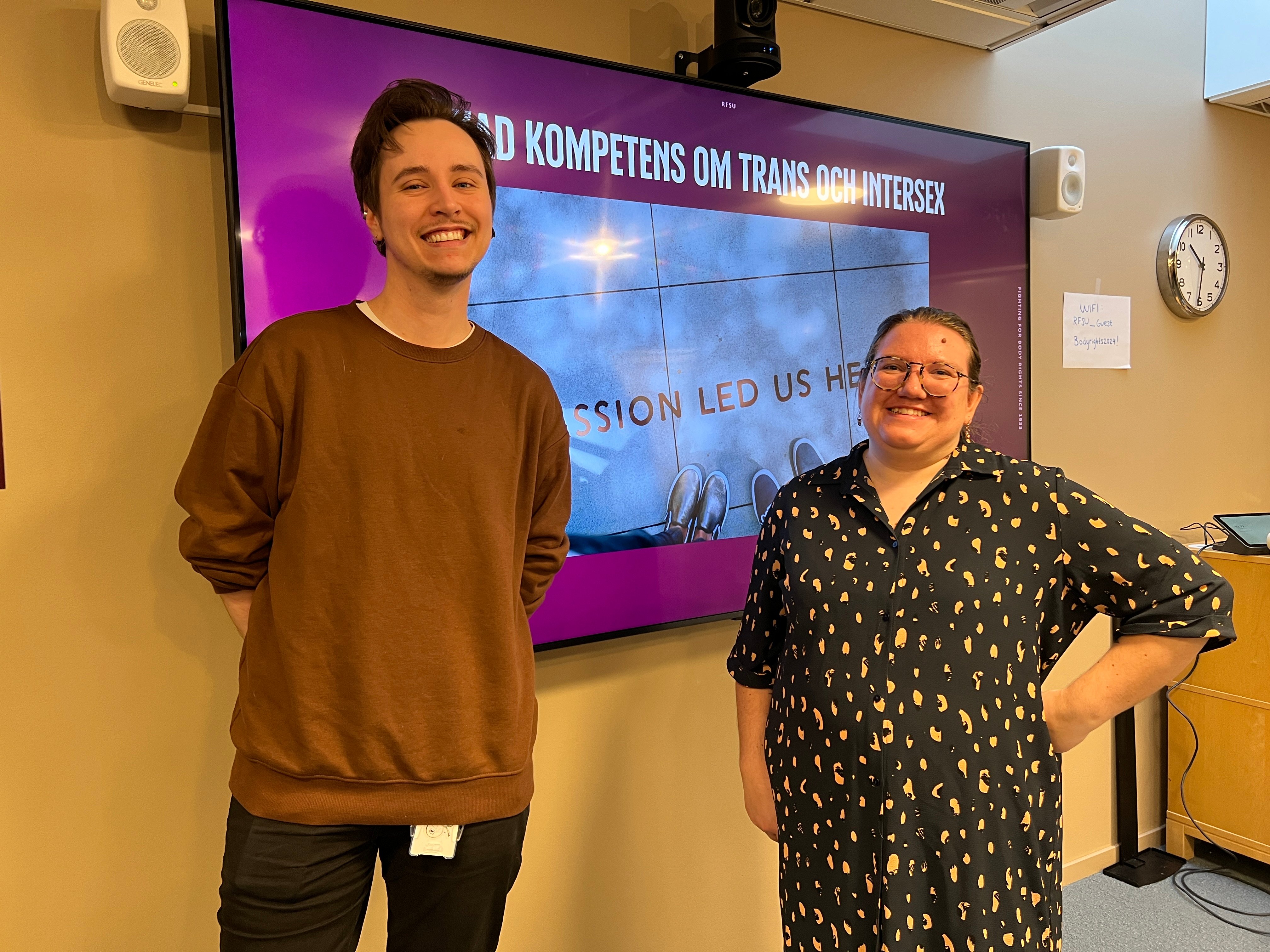 Två personer ler framför en digital skärm med texten ”Ökad kompetens om trans och intersex”.