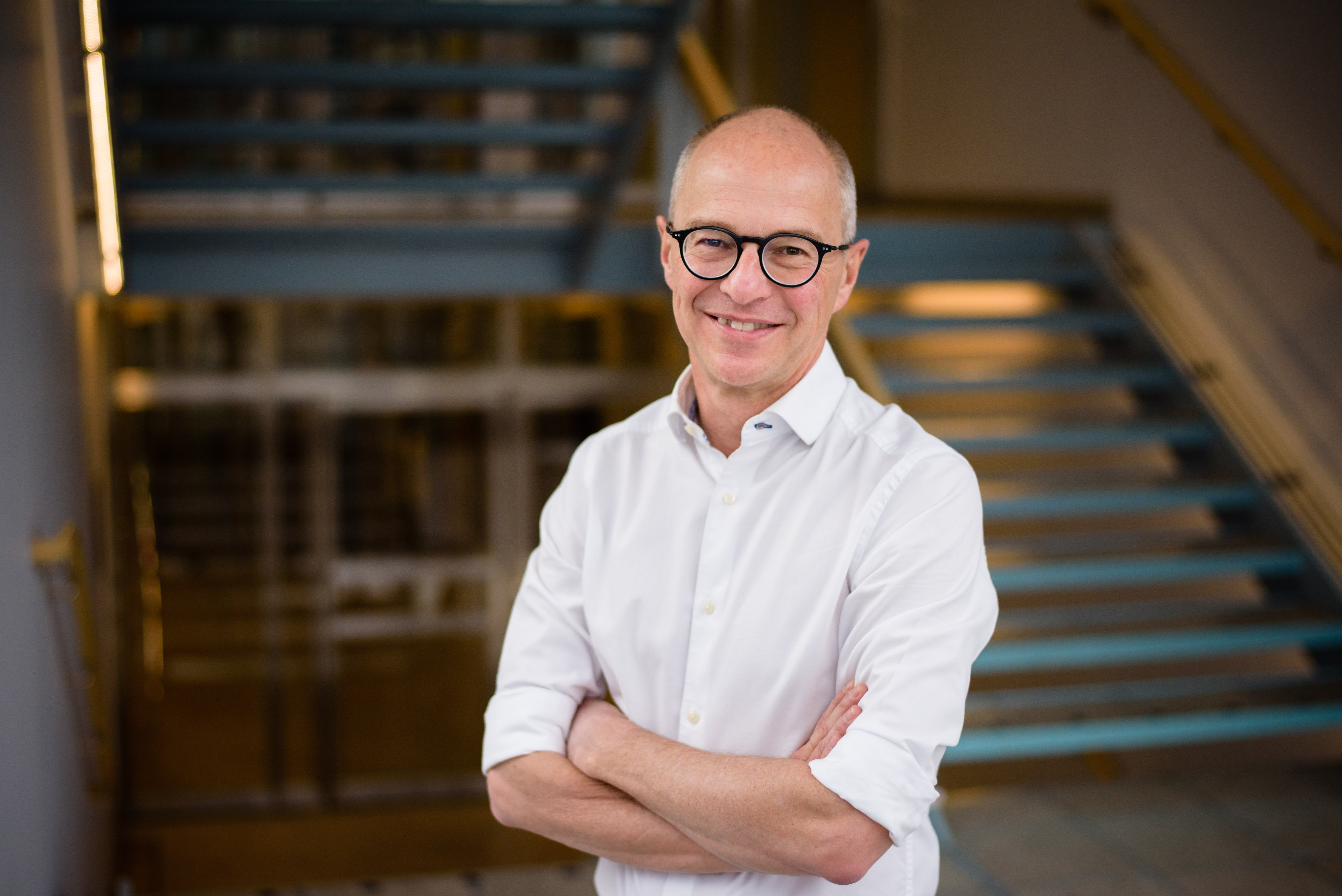 A smiling man wearing black-rimmed glasses and a white button-down shirt stands with his arms crossed. A modern indoor staircase is visible in the blurred background.