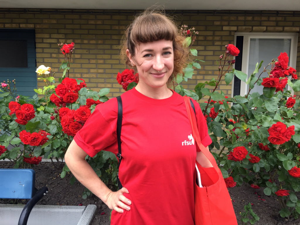 En person med ljusbrunt hår står med en röd RFSU t-shirt framför en röd rosenbuske.