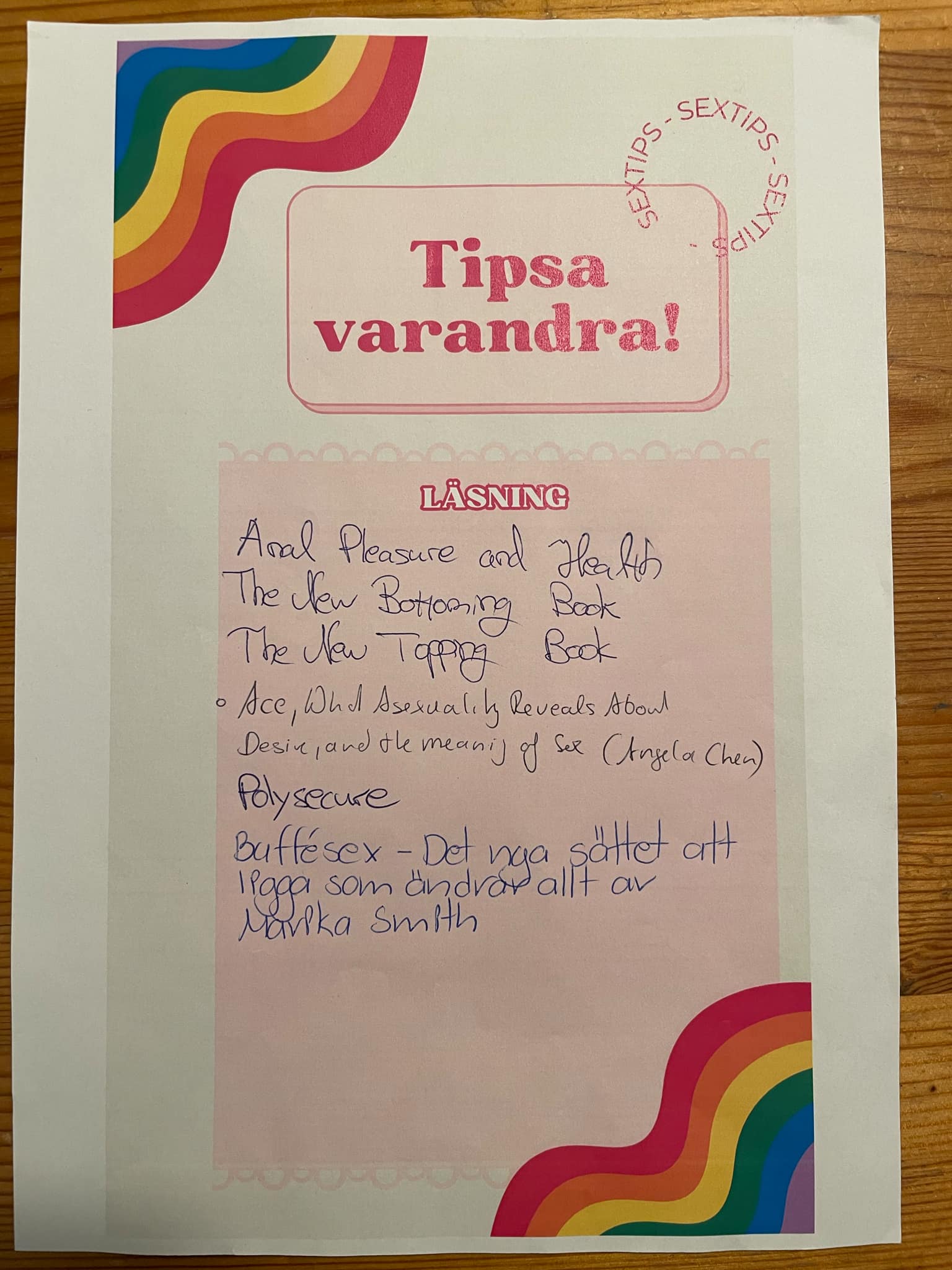 Ett papper med rubriken "Tipsa varandra!" ligger på en träyta. Bladet är dekorerat med regnbågsfärgade vågor i hörnen och en cirkulär stämpel med texten "SEXTIPS". Under rubriken "LÄSNING" finns en handskriven lista med boktips, bland annat "Polysecure", "Ace" av Angela Chen och "Buffésex" av Marika Smith.