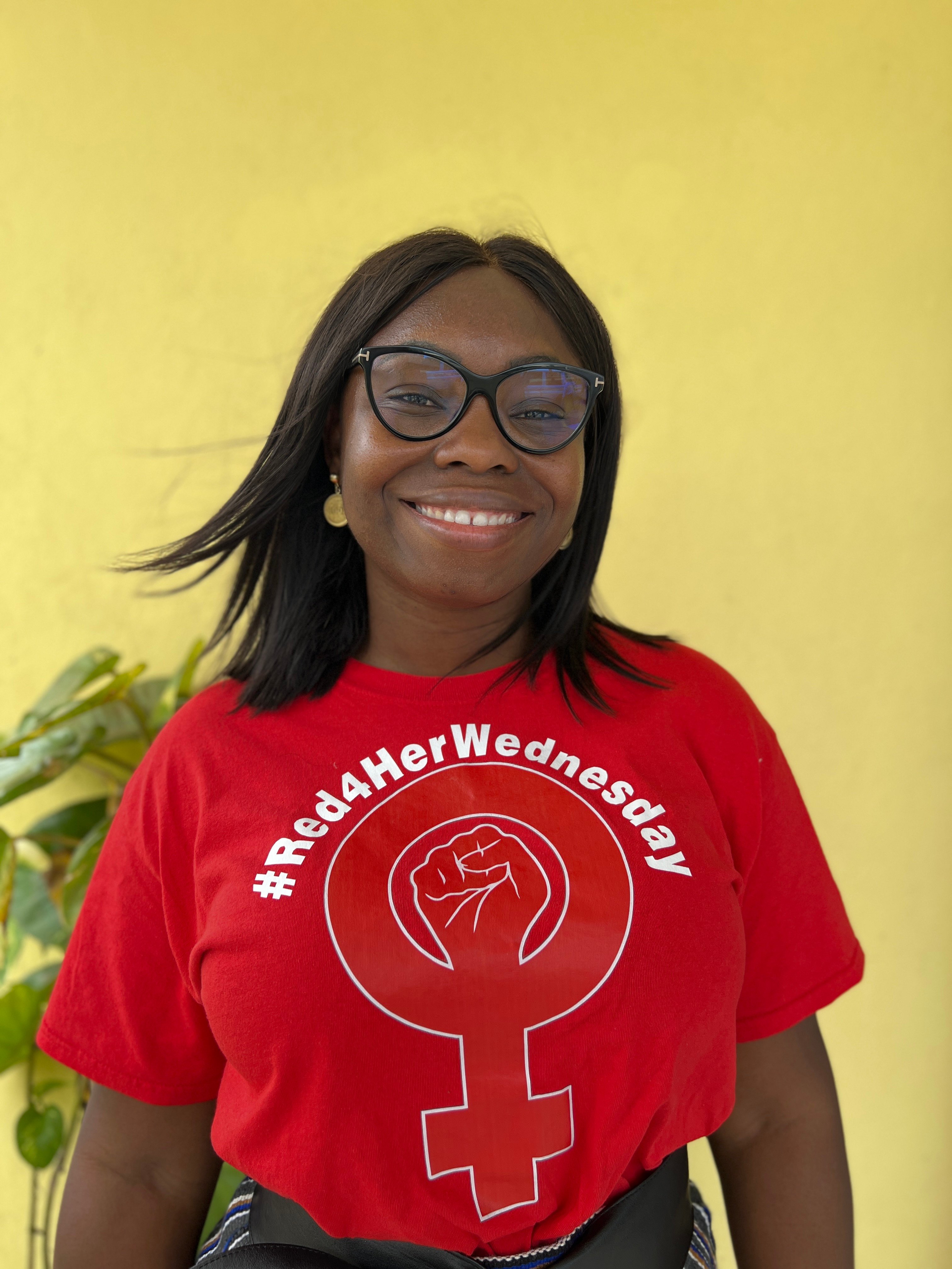 A smiling woman wearing black-rimmed glasses poses for a portrait against a plain yellow background. She wears a bright red t-shirt featuring a female symbol with a raised fist inside it and the text "#Red4HerWednesday".