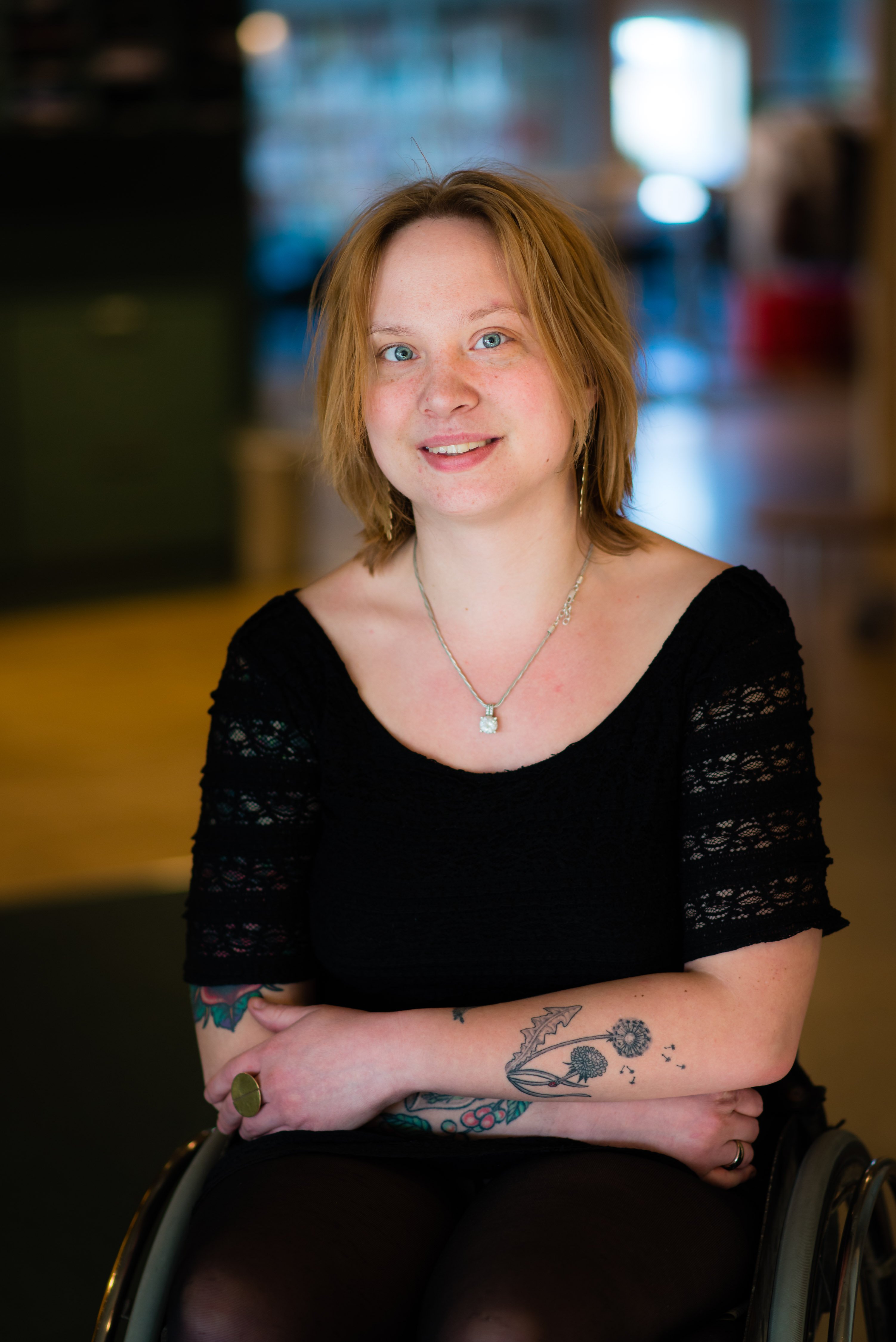 En person med blåa ögon och honungsfärgat hår sitter på sin rullstol och tittar in i kameran och småler.