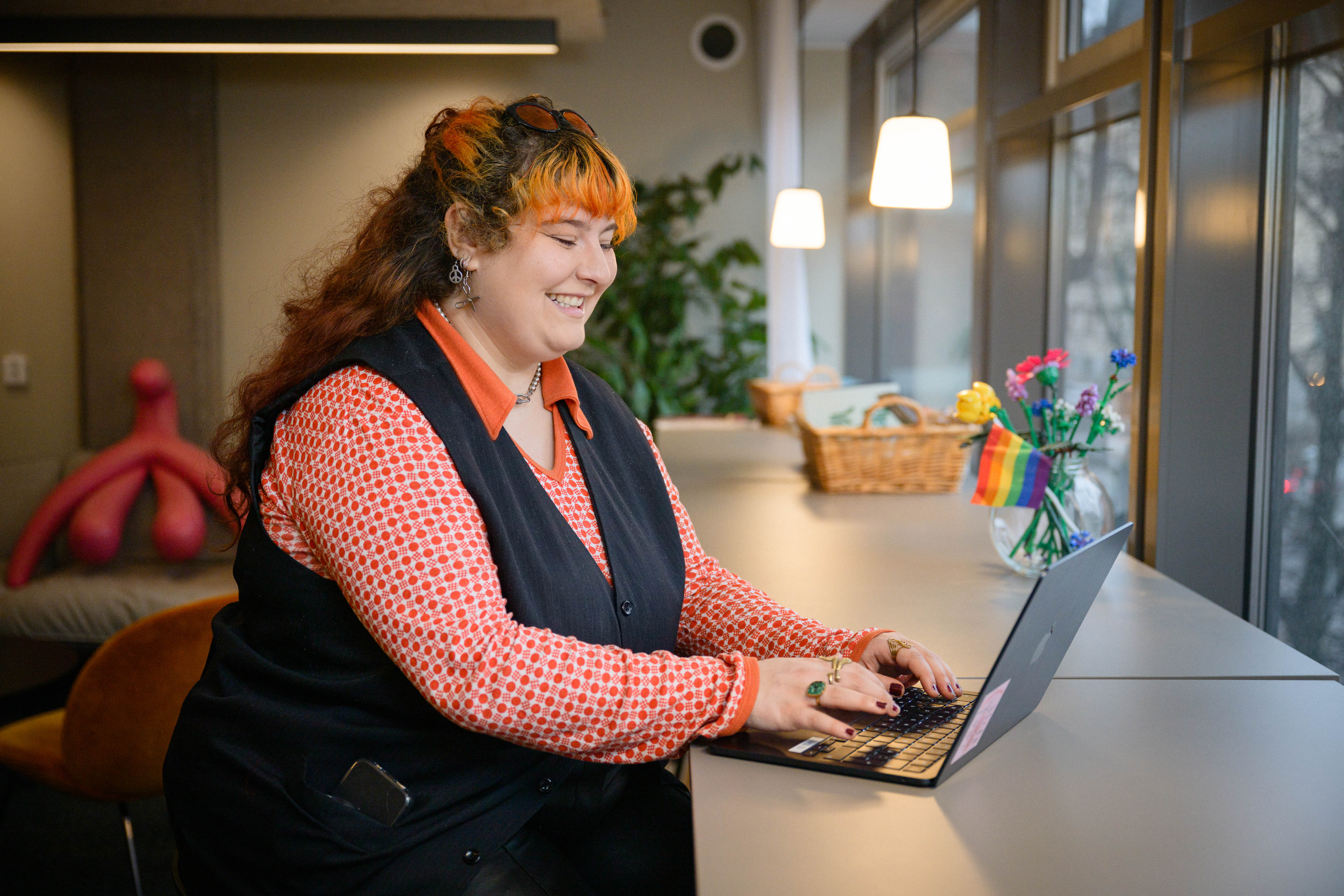 En person sitter och jobbar på en laptop och har svart väst och orangemönstrad blus