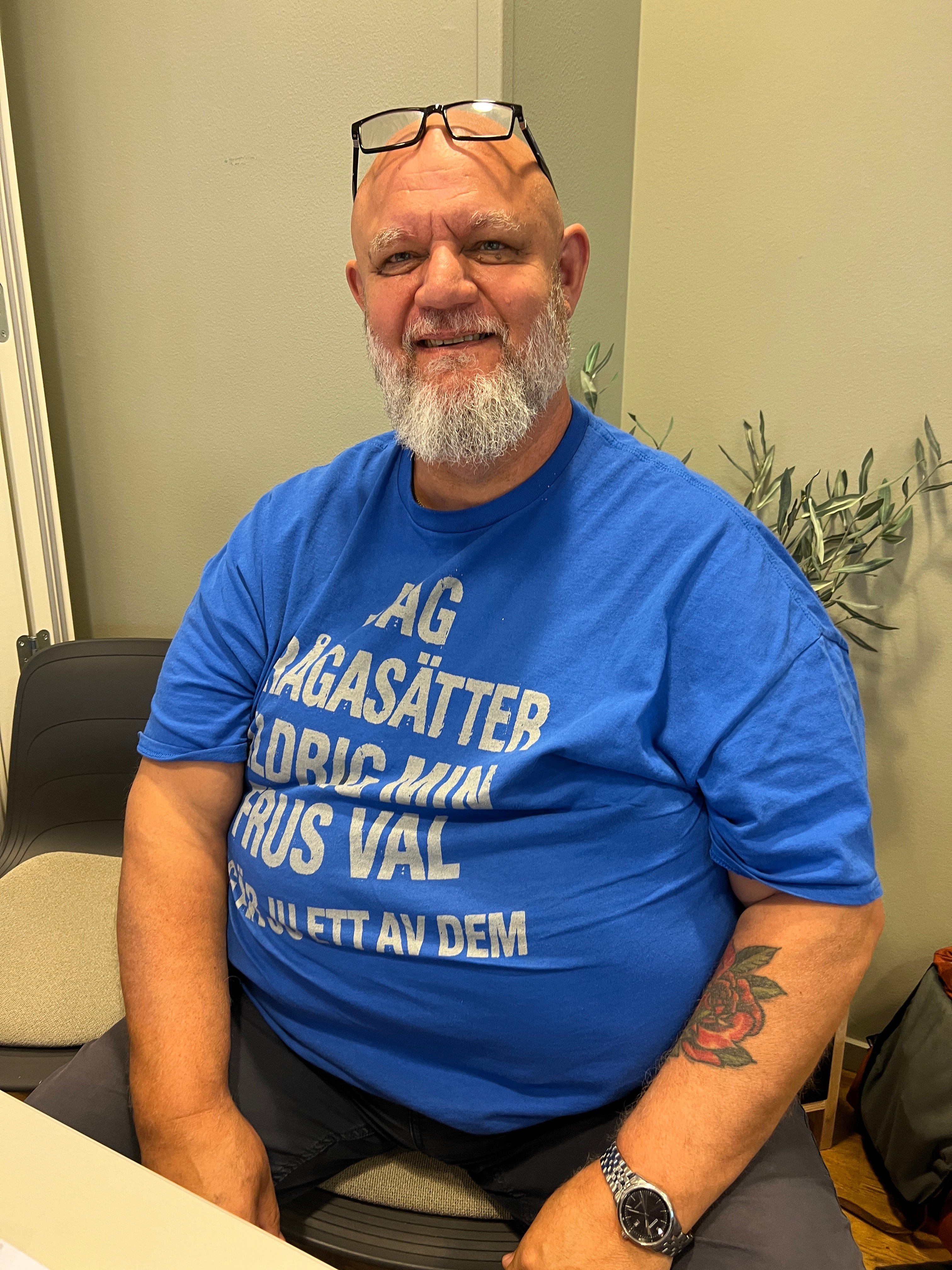 En man med blå t-shirt och glasögon på huvudet sitter på en stol och ler smått mot kameran.
