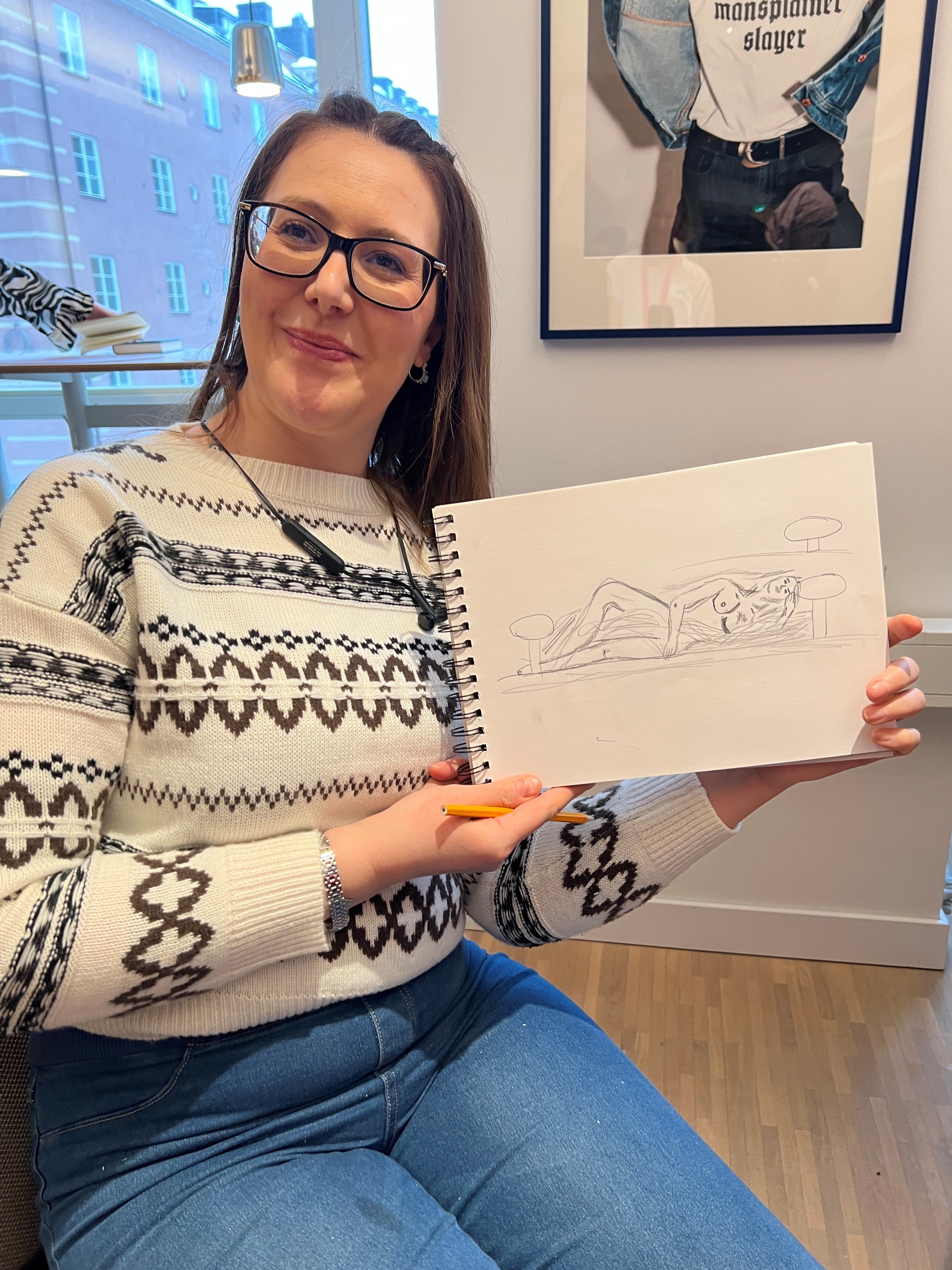 En kvinna i mönstrad tröja visar upp en detaljerad blyertsskiss av en liggande person.