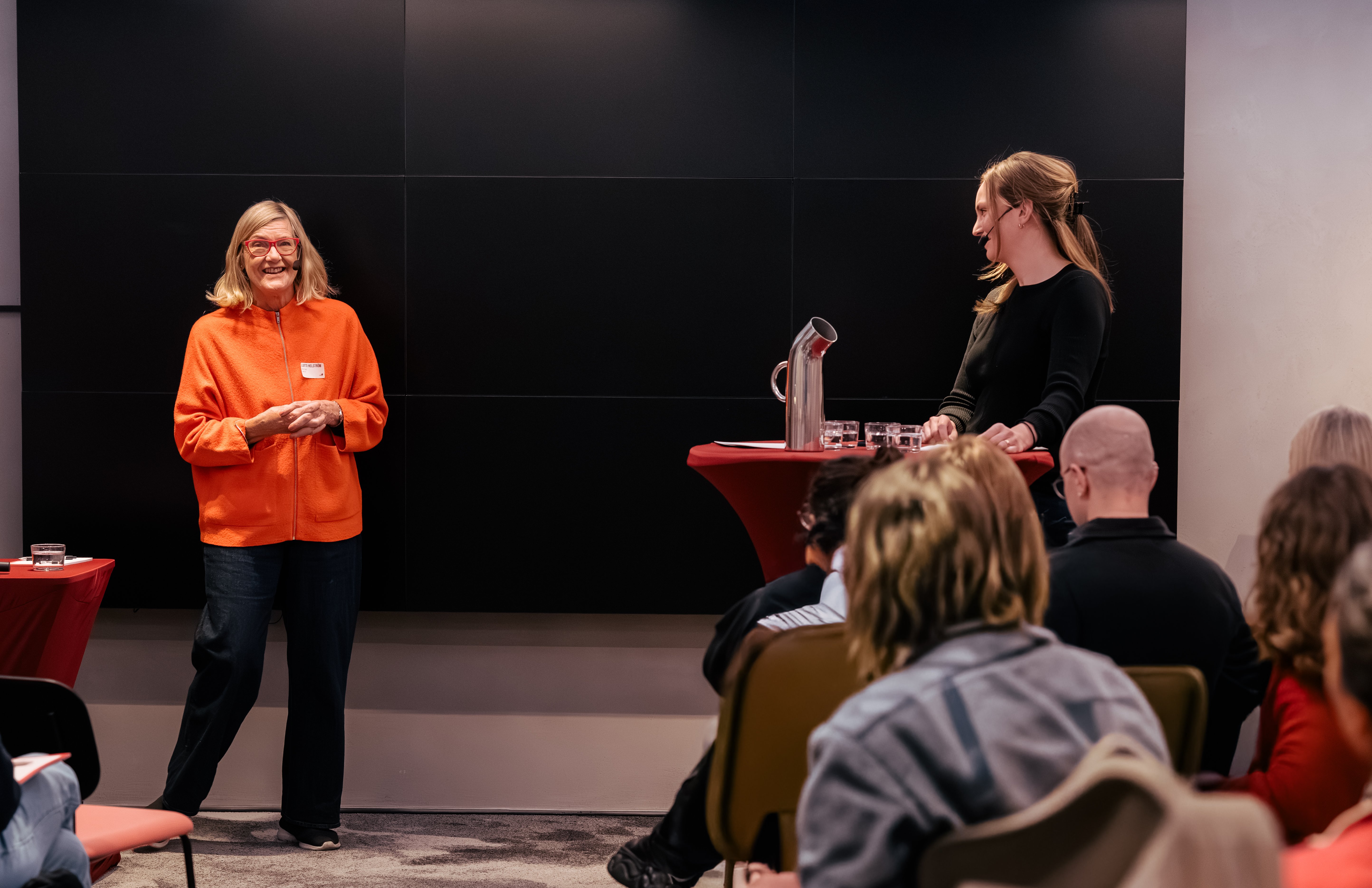 Två kvinnor står inför en publik med handfria mikrofoner och pratar