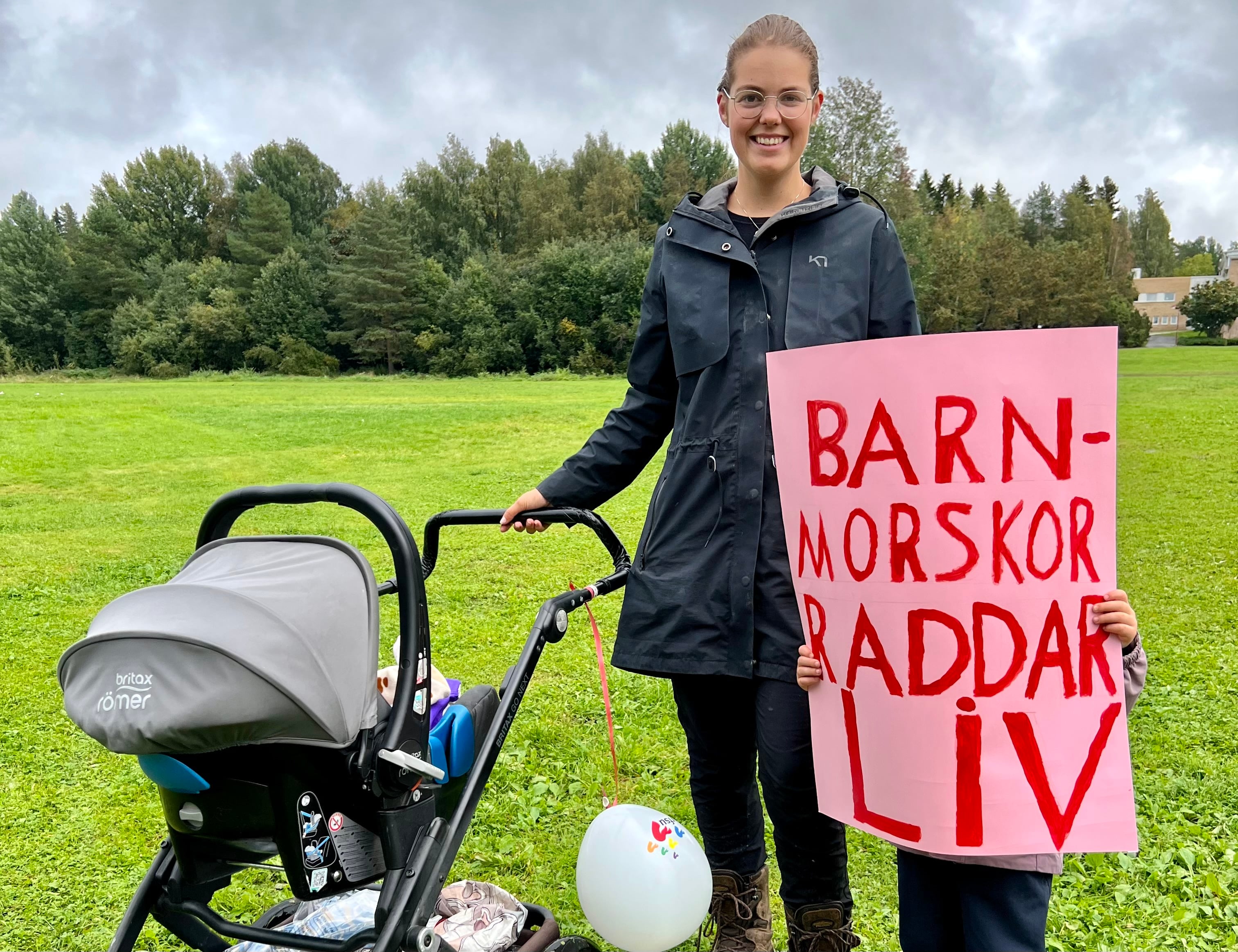 Person med glasögon står bredvid en barnvagn. Ett litet barn håller upp en skylt som det står Barnmorskor räddar liv på. 