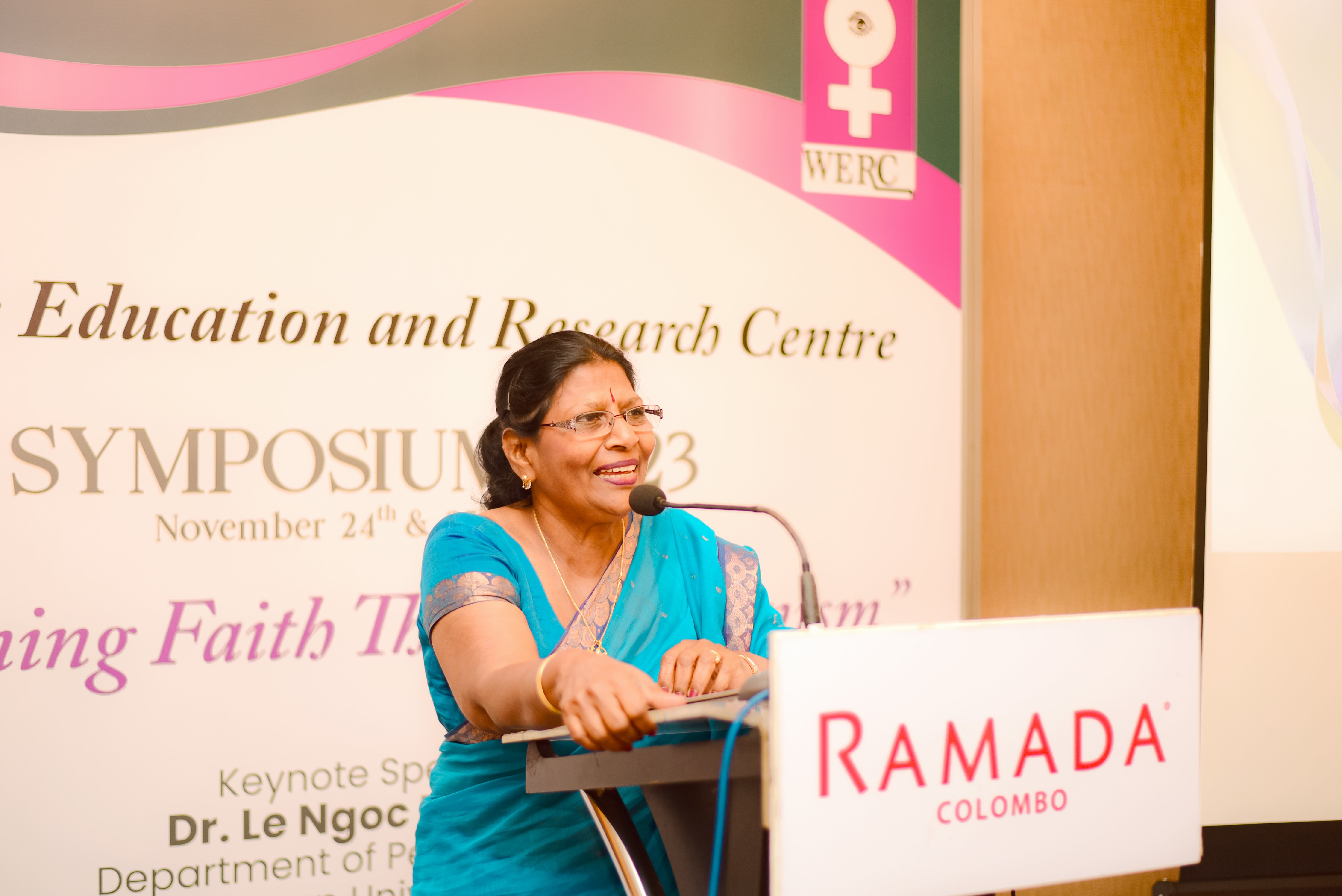 A woman in a bright blue sari speaks into a microphone at a podium branded "Ramada Colombo" during a symposium. A large background banner features the WERC logo and event text.