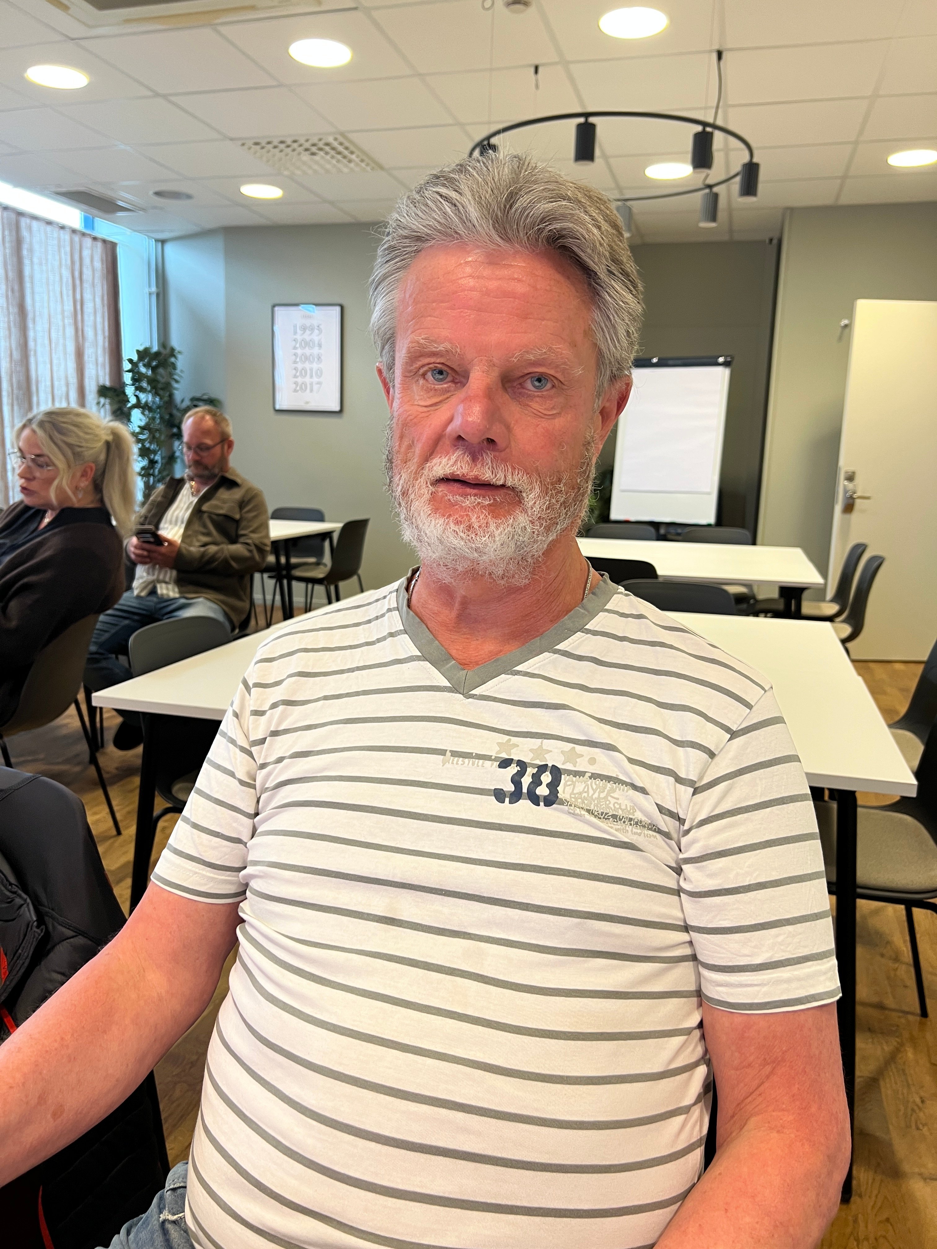 En man i vit och grå-randig t-shirt sitter i ett rum med bord och stolar bakom och tittar mot kameran.