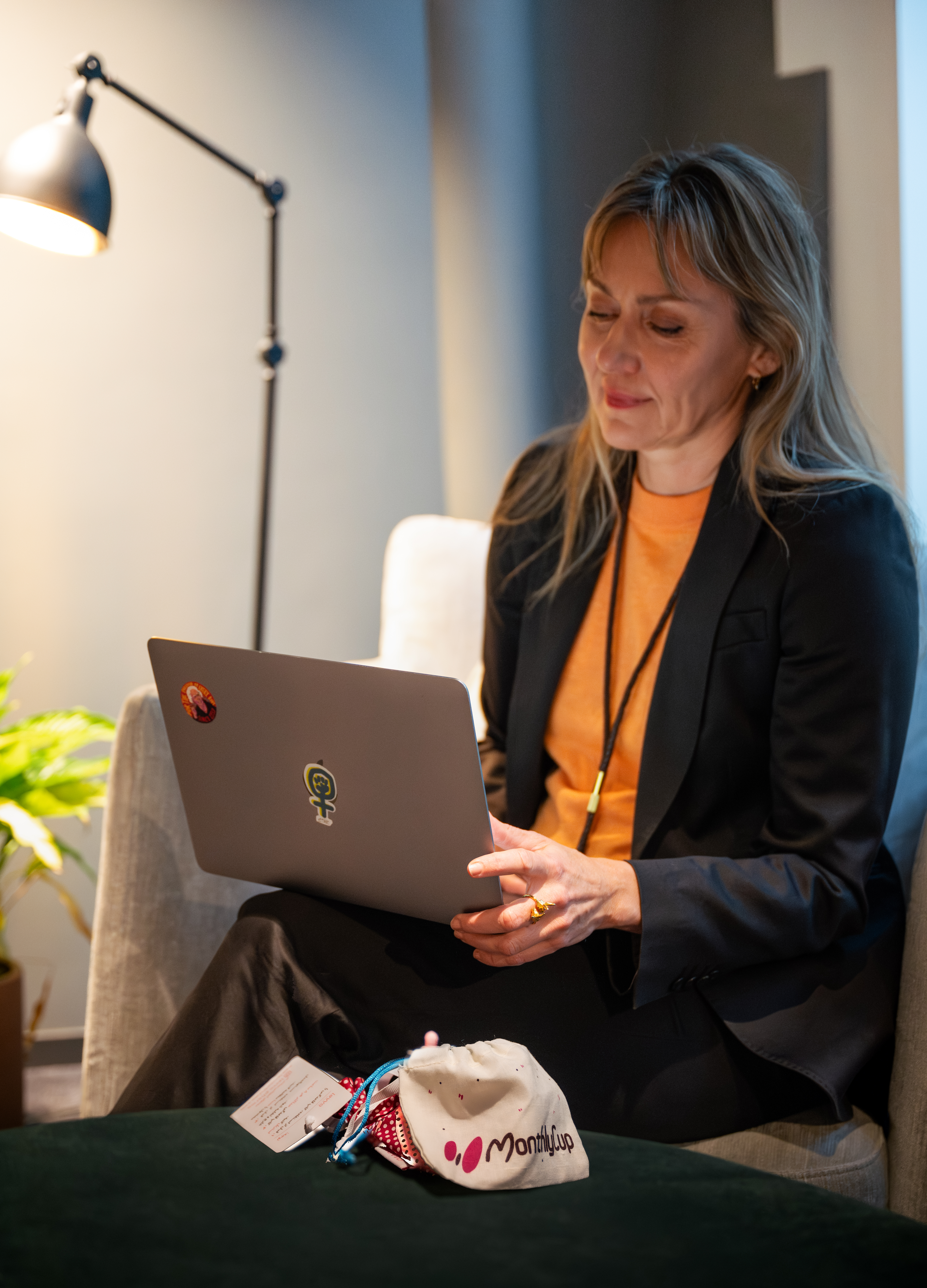 Person i mörk kavaj och orange tröja sitter i en fåtölj med en bärbar dator i knät. På bordet framför ligger en liten tygpåse med texten ”MonthlyCup”.
