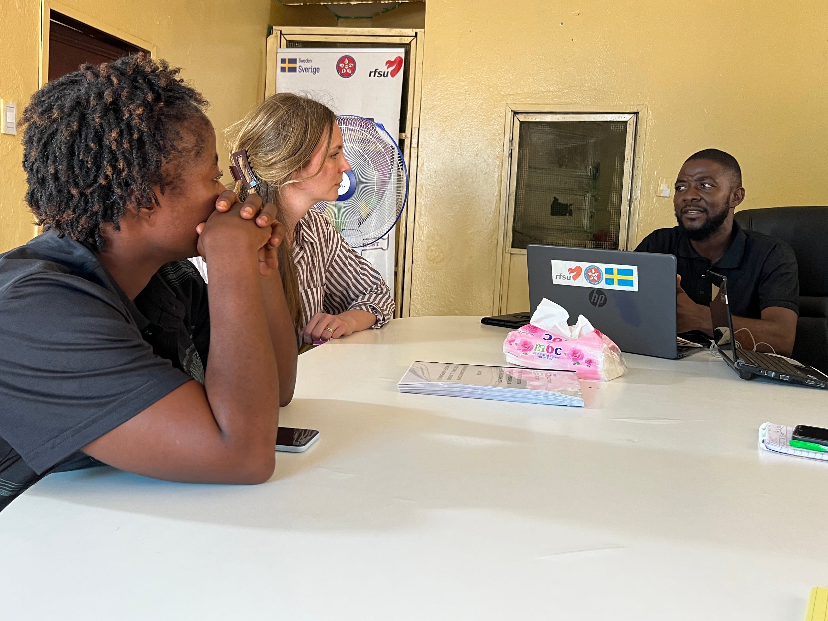 A man sits at a table speaking to two women who are listening attentively. A laptop with stickers, including a Swedish flag and an RFSU logo, rests on the table alongside a pink tissue box.