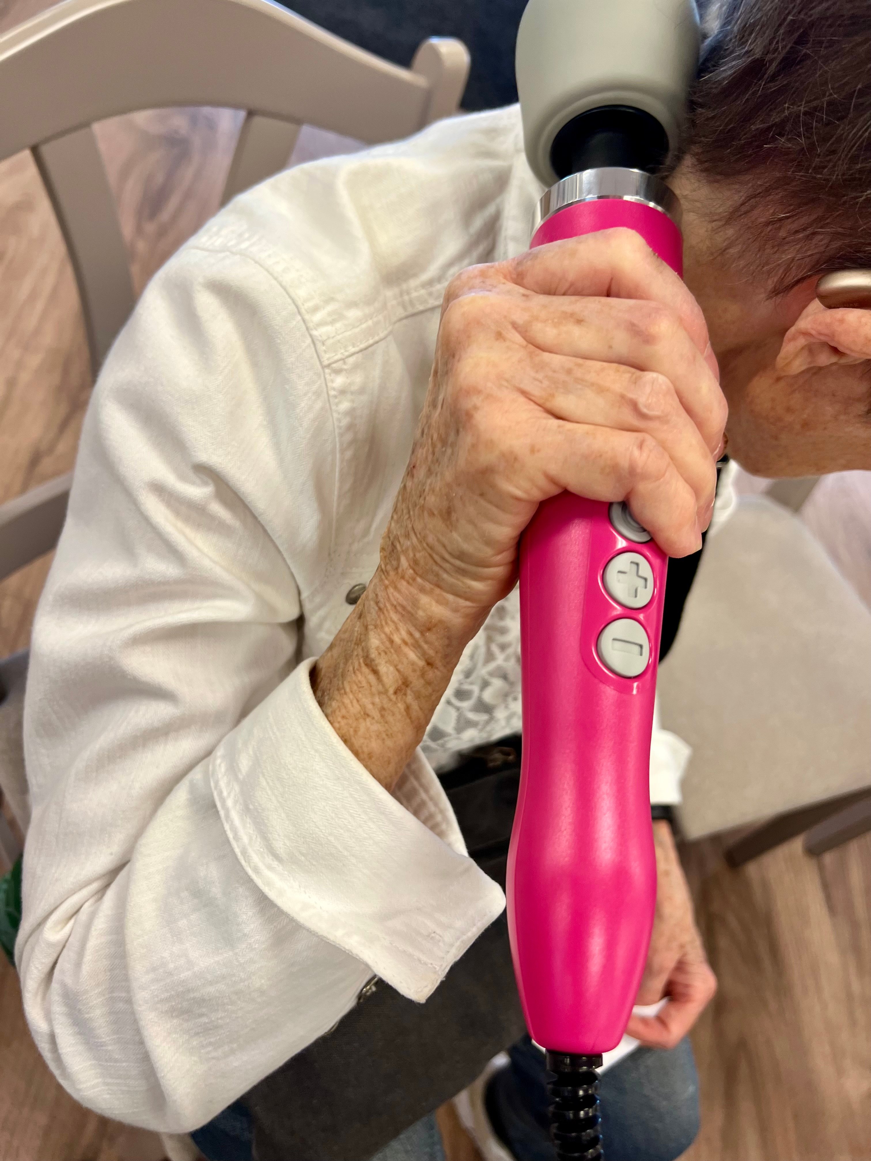 En hand håller en rosa massageapparat mot nacken på en person i vit jeansjacka.
