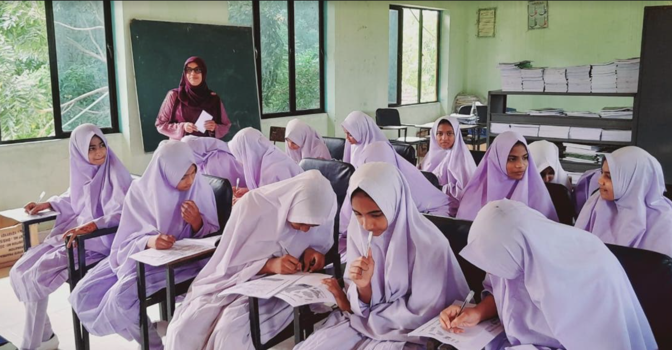A classroom full of female students wearing matching light purple hijabs sit at their desks, focused on writing in their notebooks. A teacher wearing a dark maroon hijab stands at the front of the room near a blackboard.