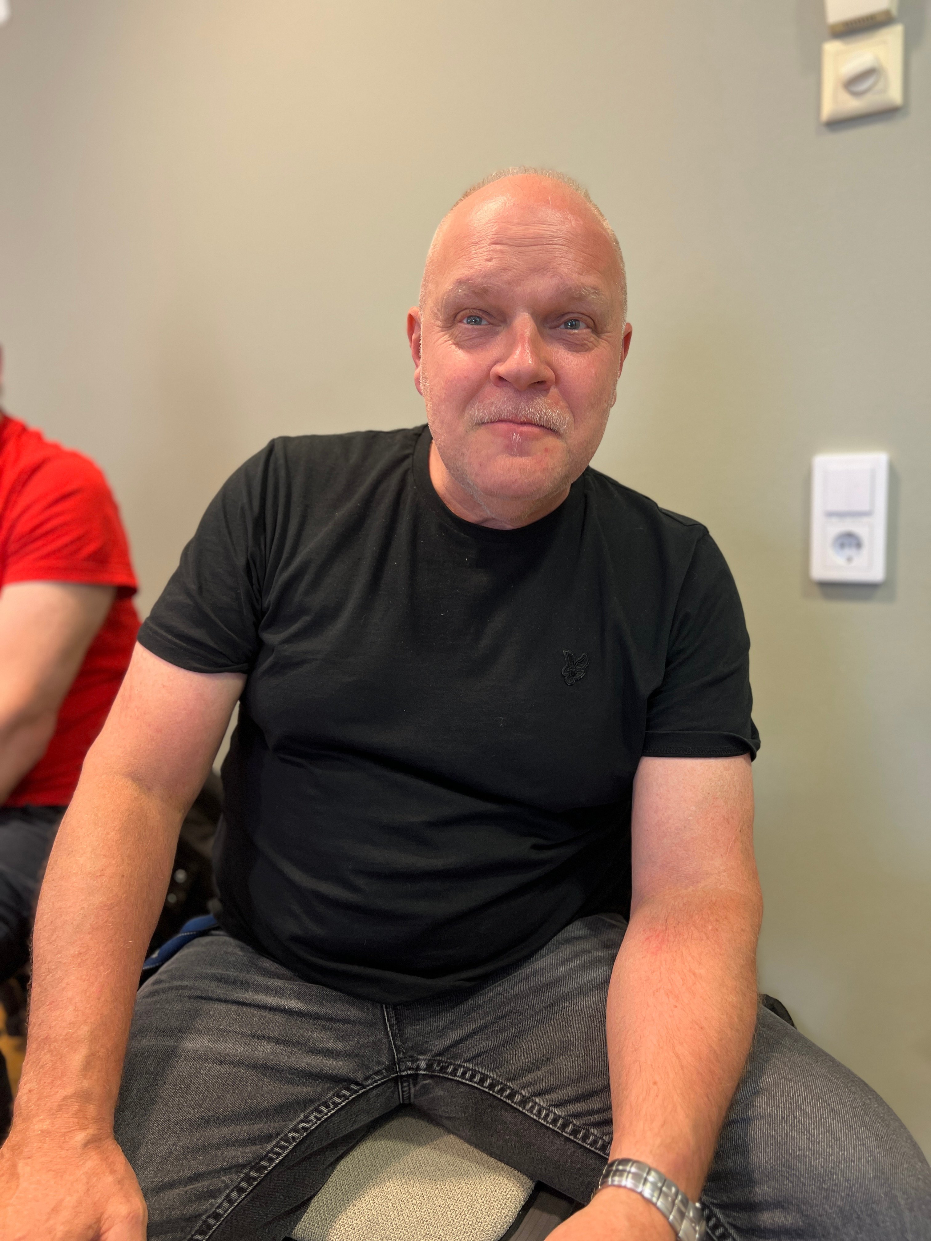 En man i svart t-shirt sitter framför en grå vägg och ler smått mot kameran.
