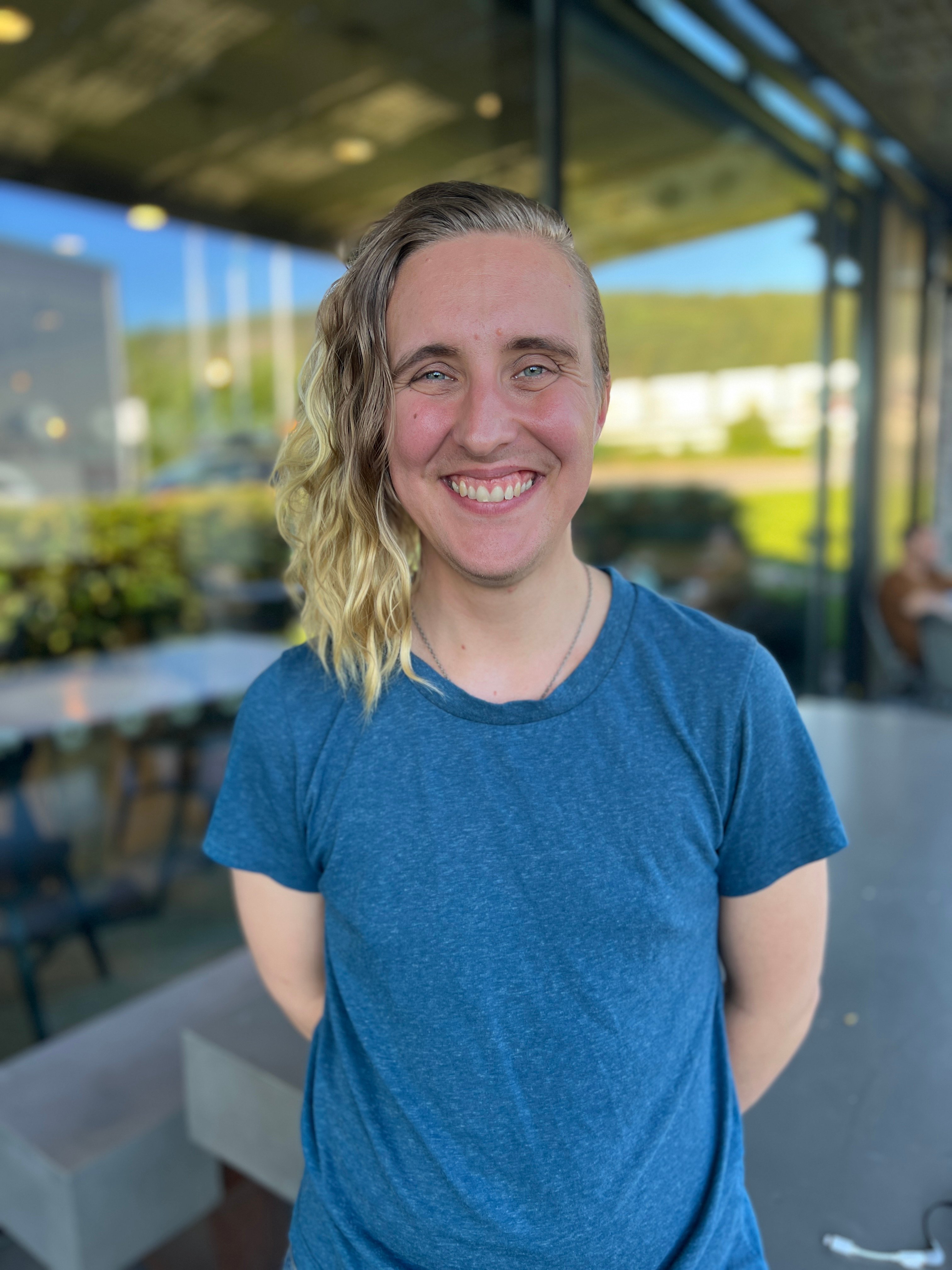Leende person i blå t-shirt med blont, vågigt hår står utomhus framför en glasfasad.