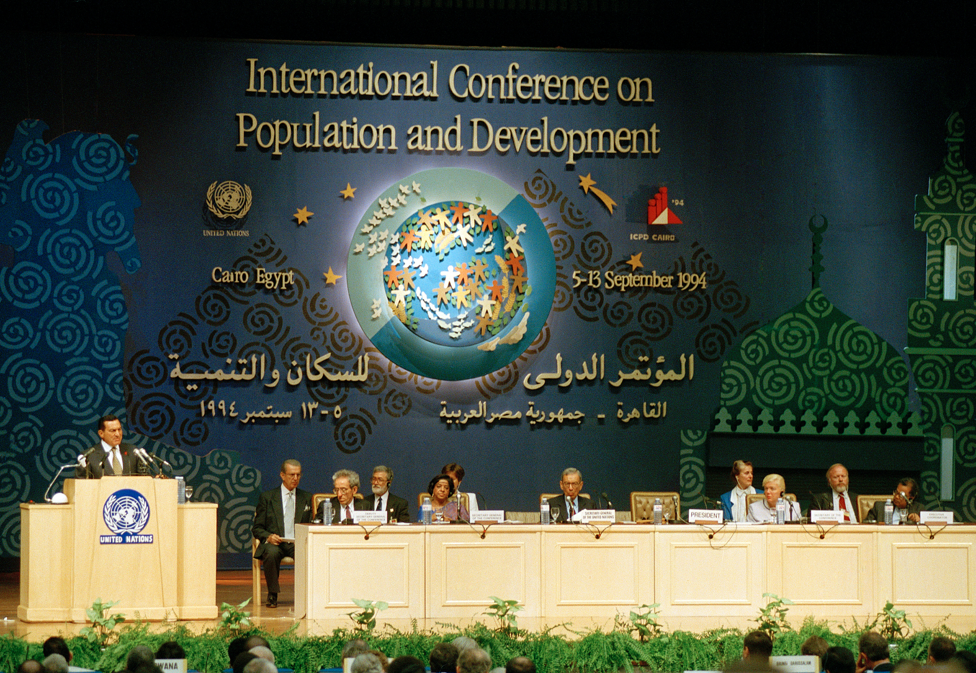 A speaker addresses an audience from a United Nations podium during the 1994 International Conference on Population and Development in Cairo, Egypt. A panel of officials is seated at a long table in front of a large, decorative blue backdrop.