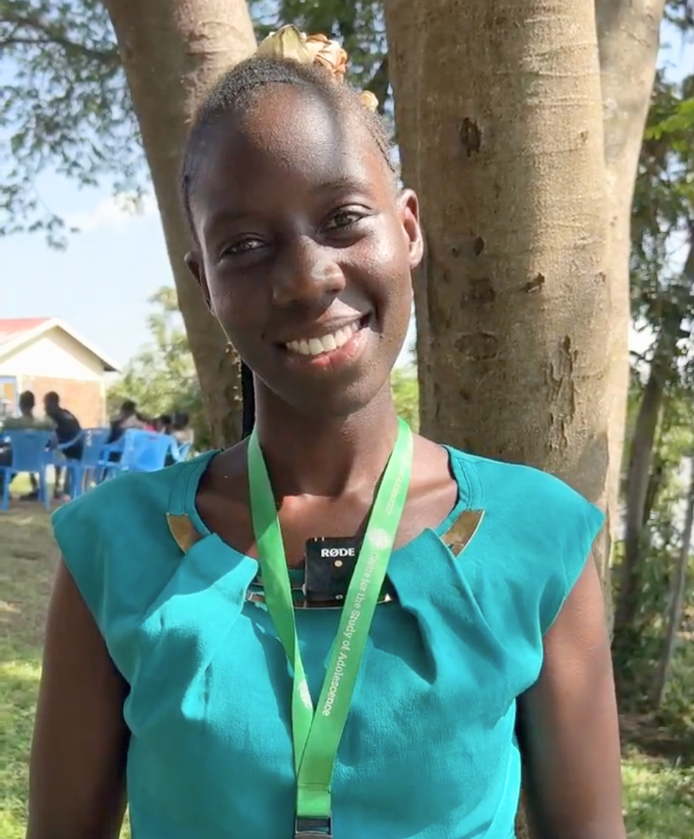 A smiling woman stands outdoors in front of a tree, wearing a teal dress and a green lanyard. A small wireless microphone is clipped to the center of her neckline.