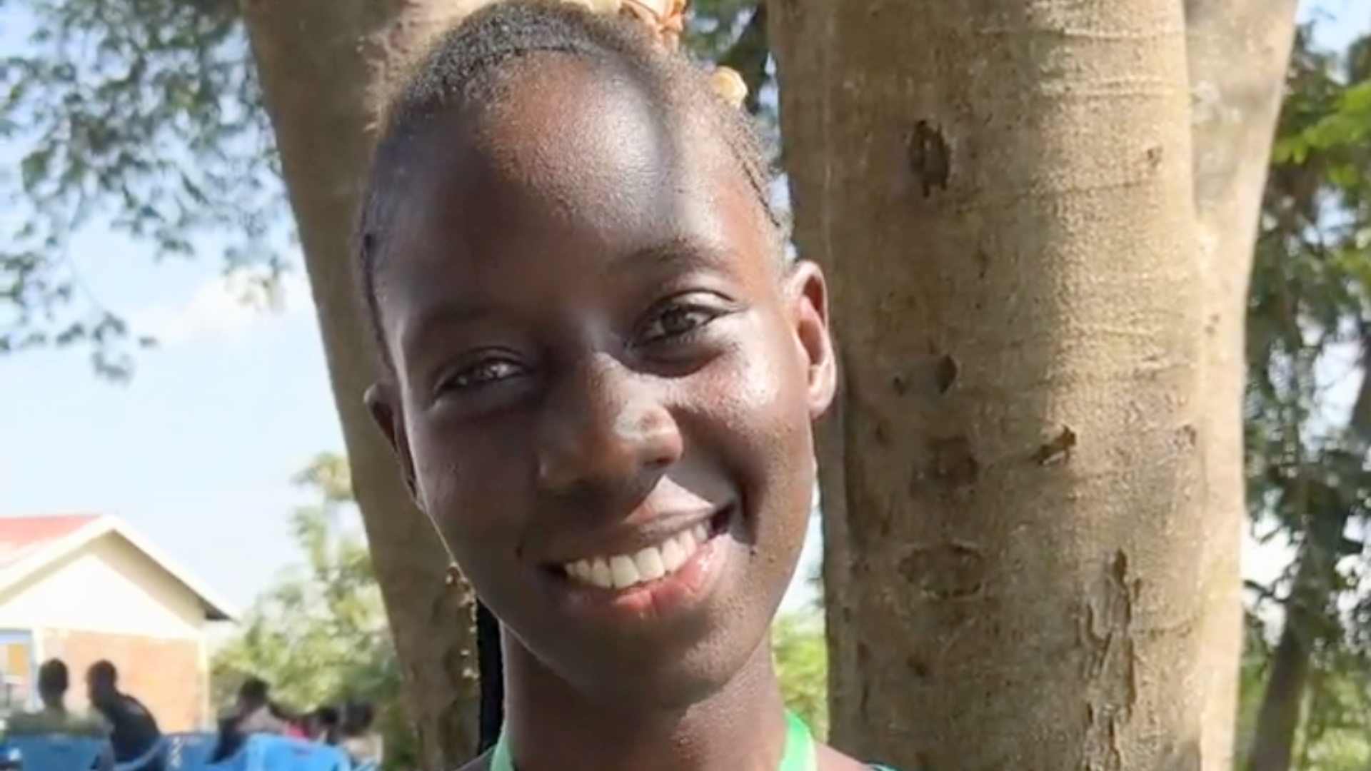 A smiling woman stands outdoors in front of a tree, wearing a teal dress and a green lanyard. A small wireless microphone is clipped to the center of her neckline.