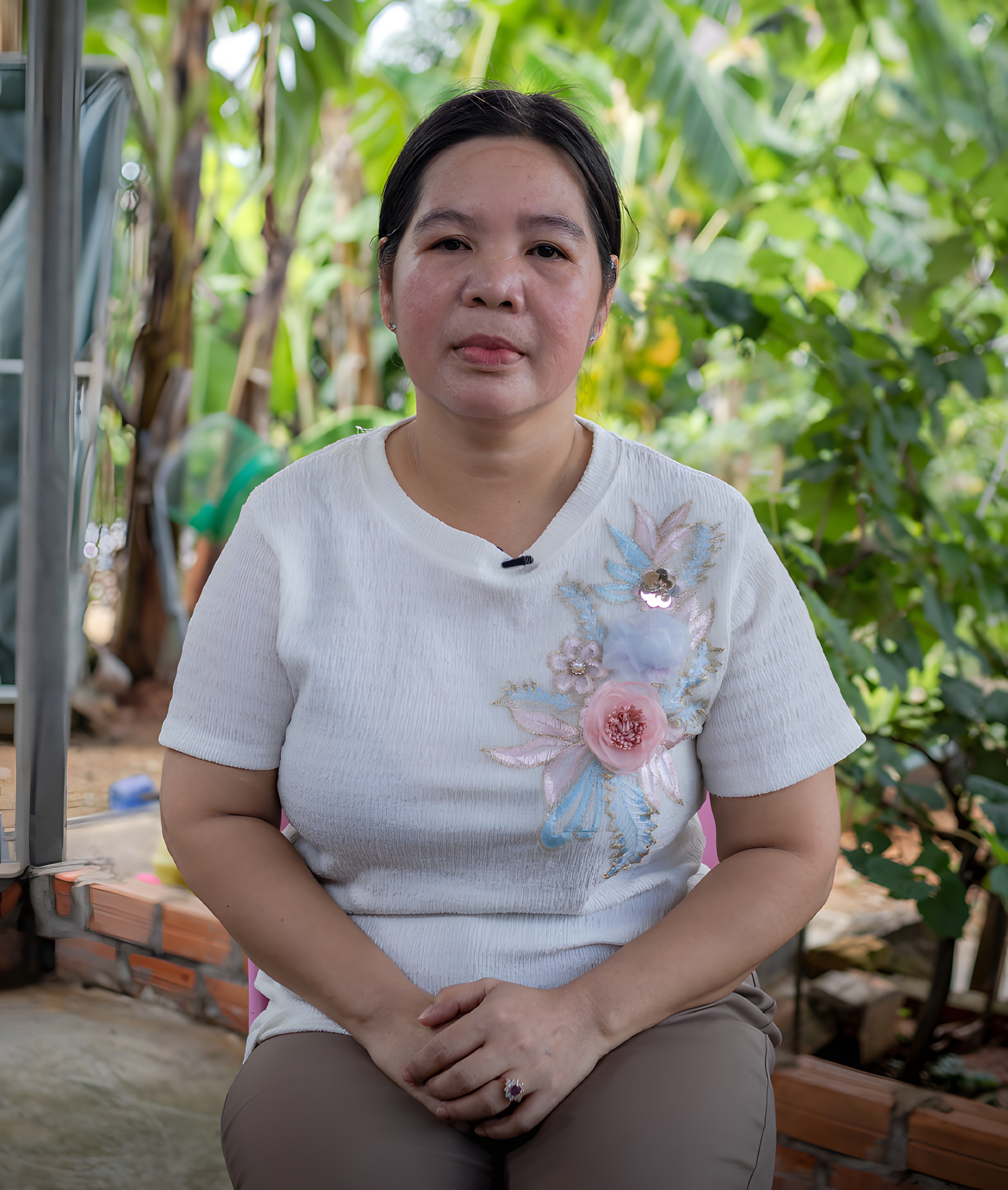 A woman with dark hair tied back sits with her hands clasped, wearing a white top adorned with embroidered pink and blue flowers. She poses against a blurred background of lush green foliage.