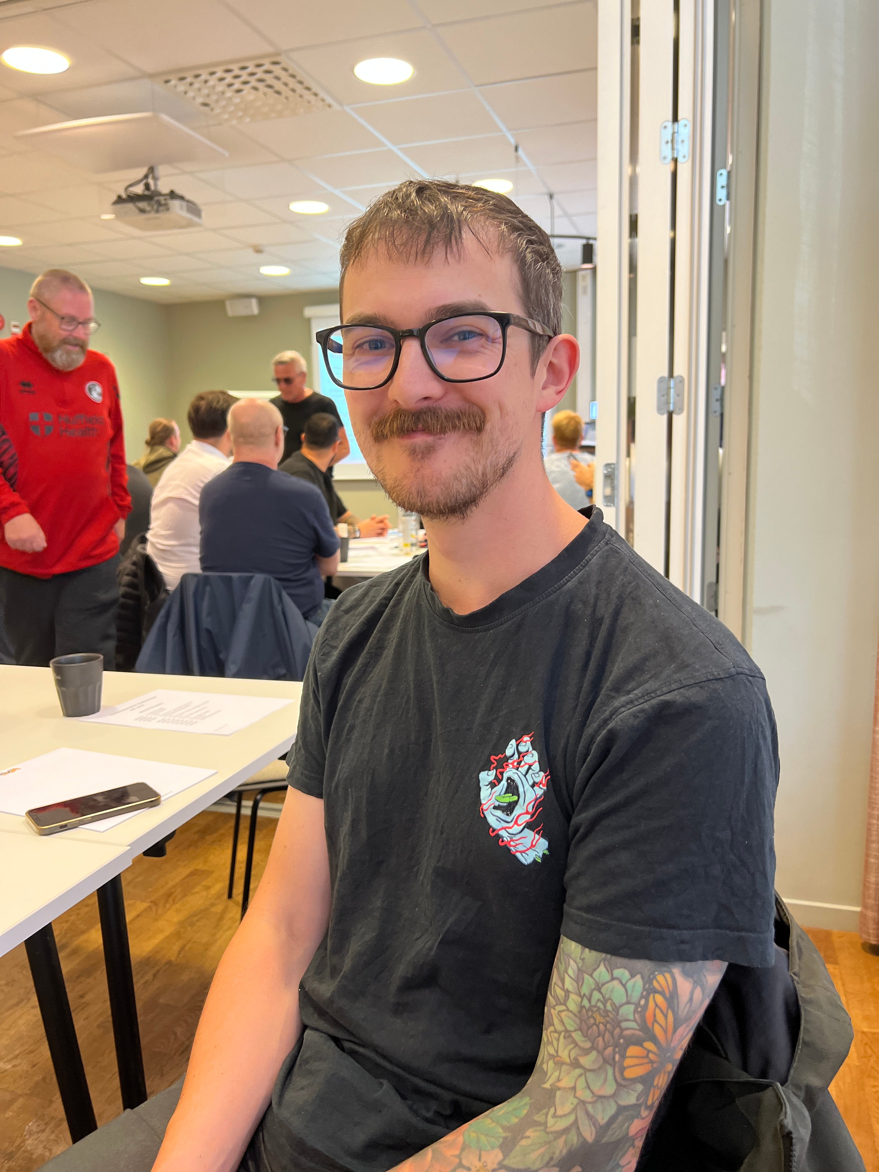 En ung man i svart t-shirt och glasögon sitter på en stol, tittar mot kameran och ler.