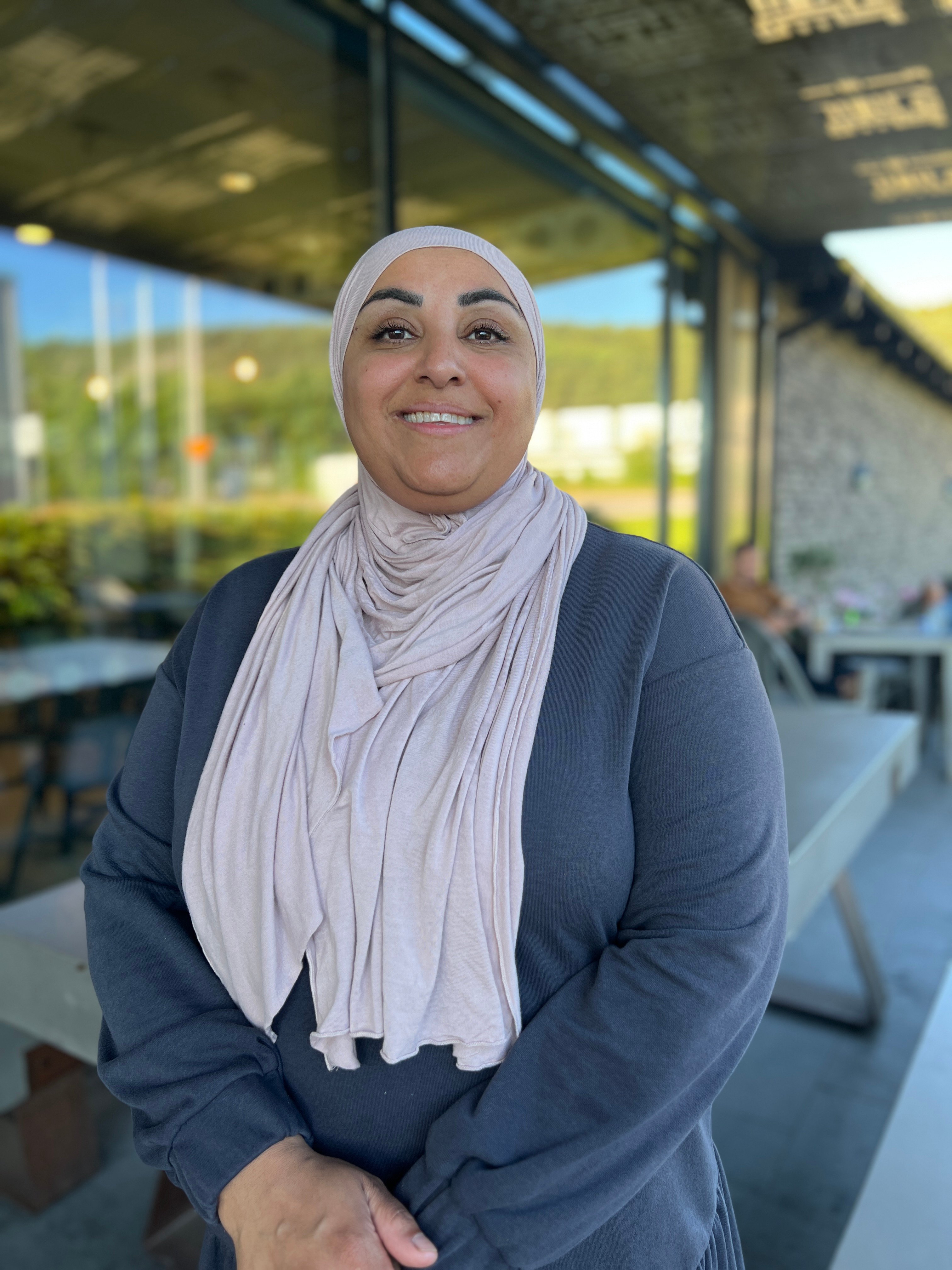 Leende person i rosa hijab och mörkblå tröja står utomhus framför en glasfasad.