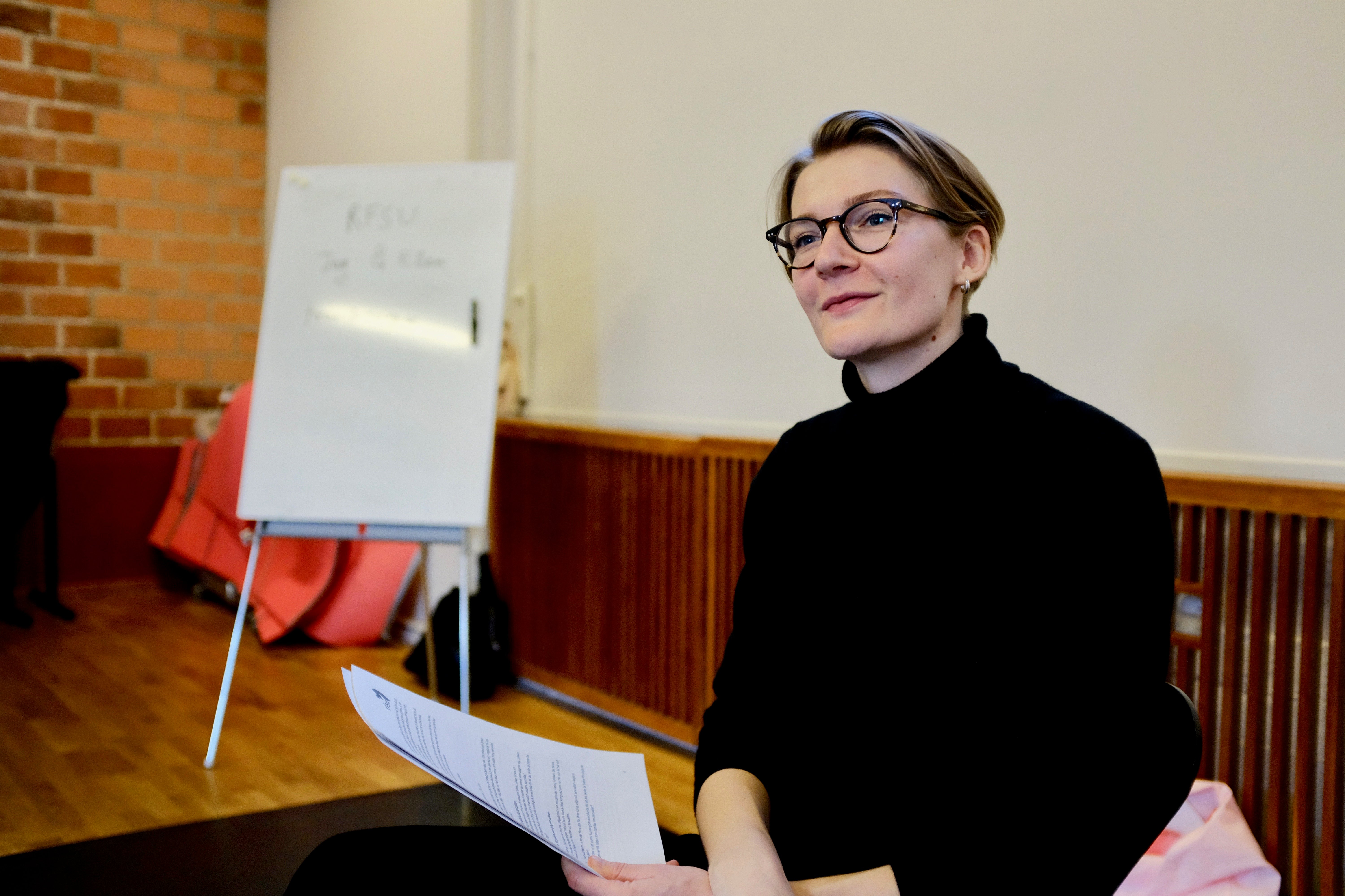 En person med kort hår och mörka glasögon sitter i en lokal och håller i några papper. Personen är klädd i en svart polotröja och tittar lite åt sidan med ett milt leende.