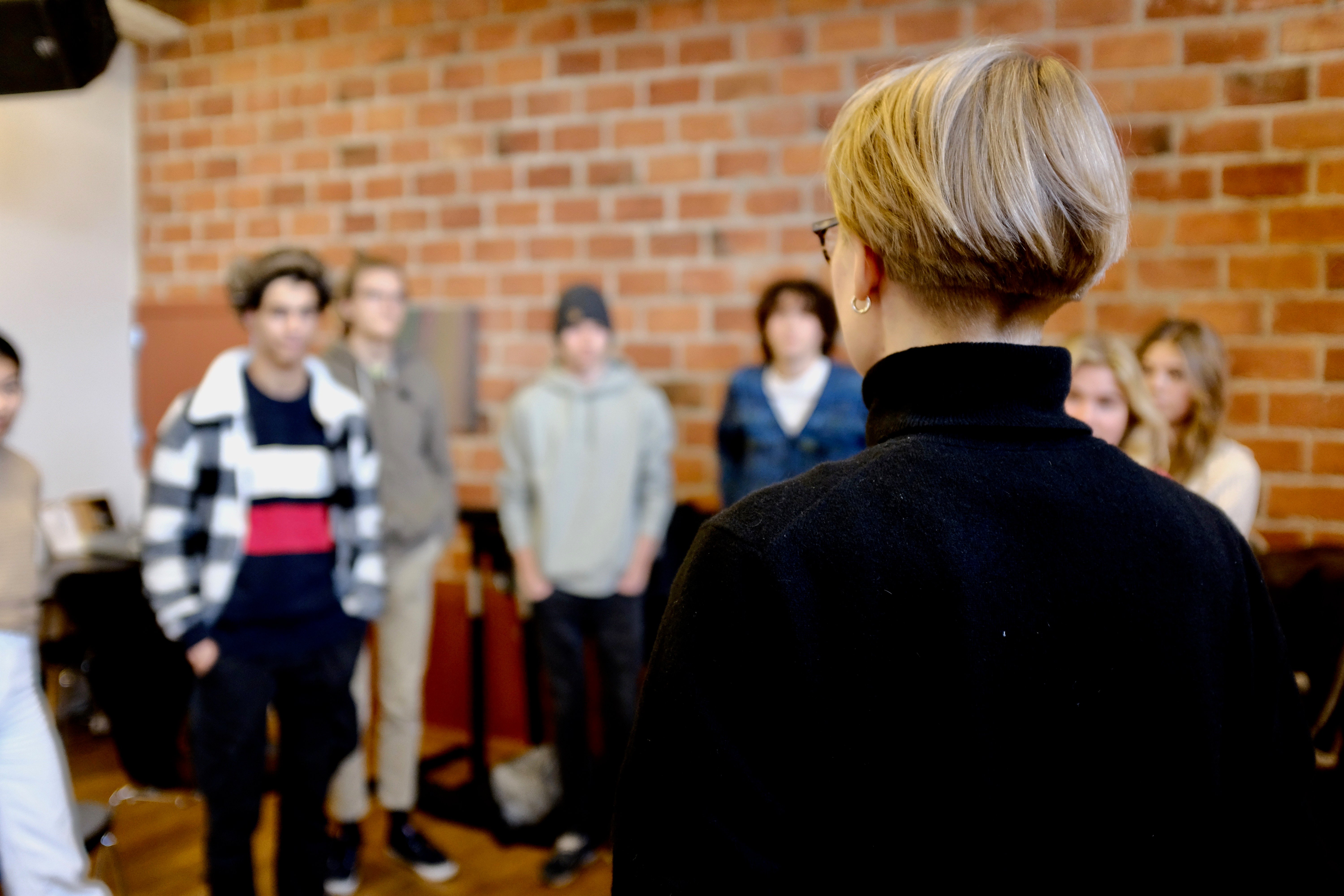 Person i svart polotröja sedd bakifrån, vänd mot en grupp ungdomar framför en tegelvägg.