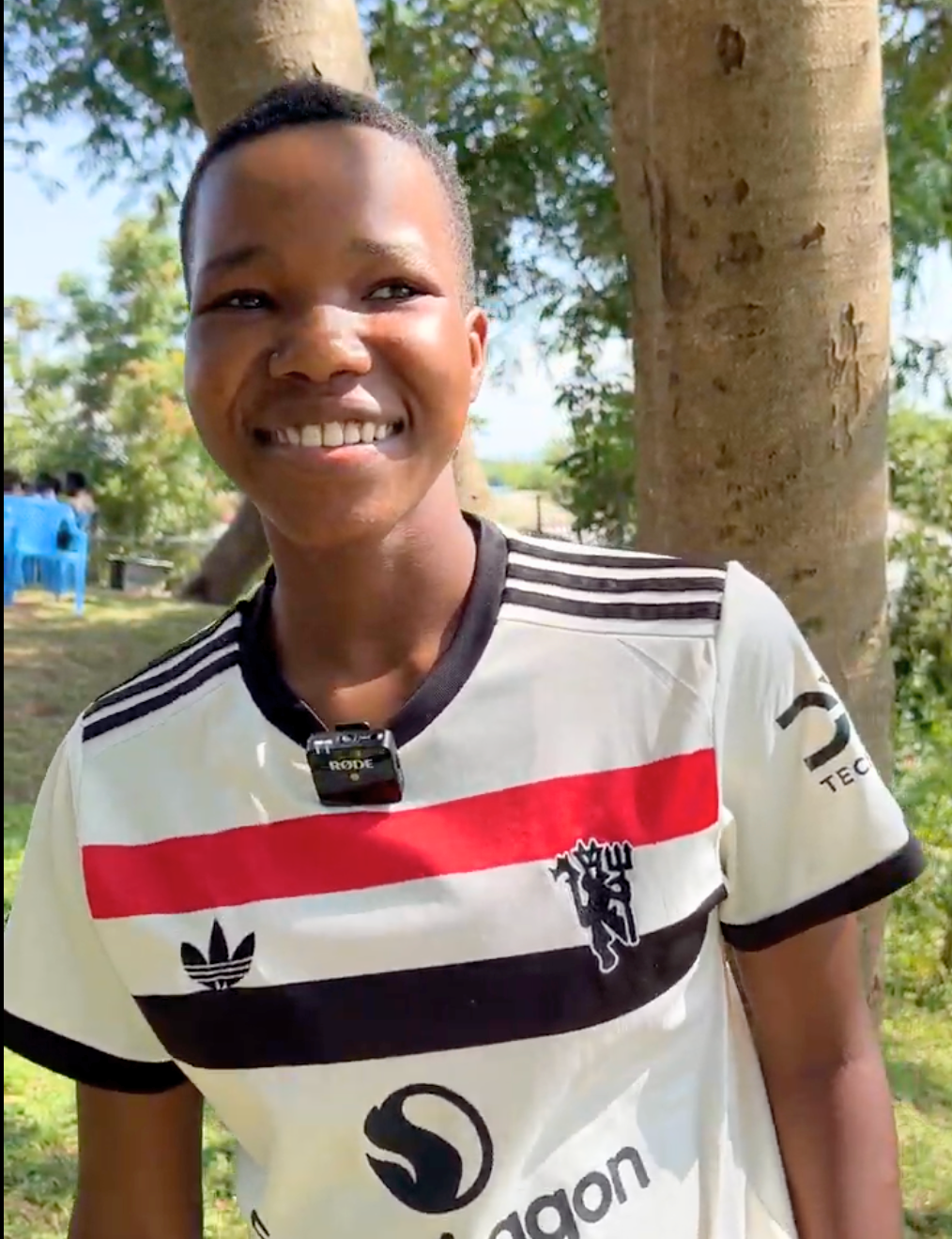 A smiling individual stands outdoors in front of trees, wearing a white sports jersey with red and black stripes. A small wireless microphone is clipped to the collar of the shirt.