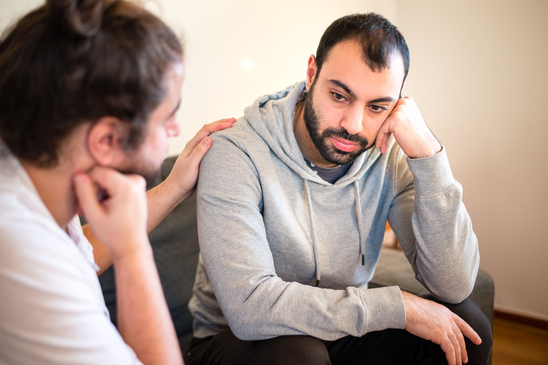 Två män sitter på en soffa. Den ena klappar den andra på axeln och verkar bekymrad