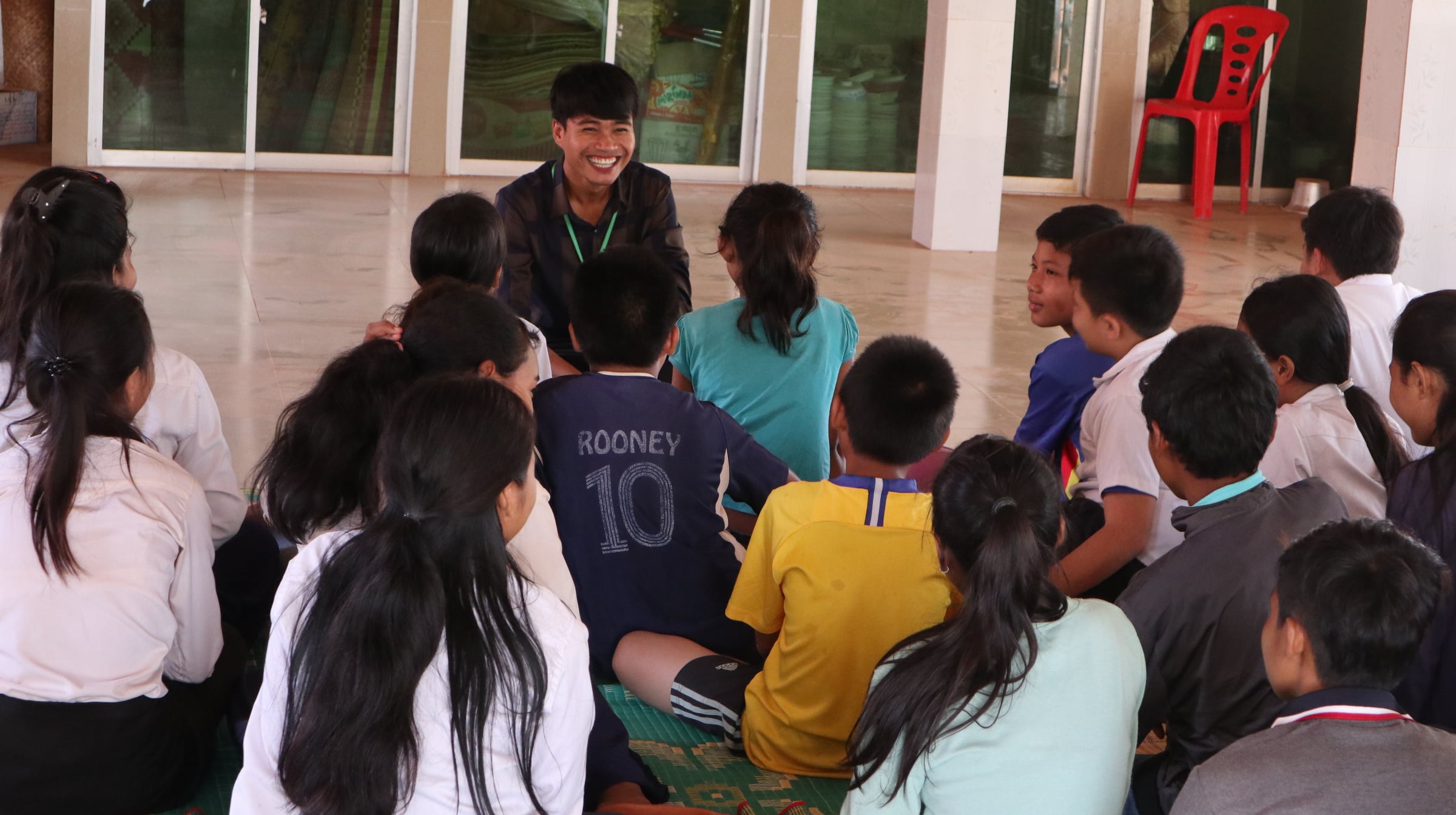 A group of children are listening to a young man. They are all sitting on the floor.