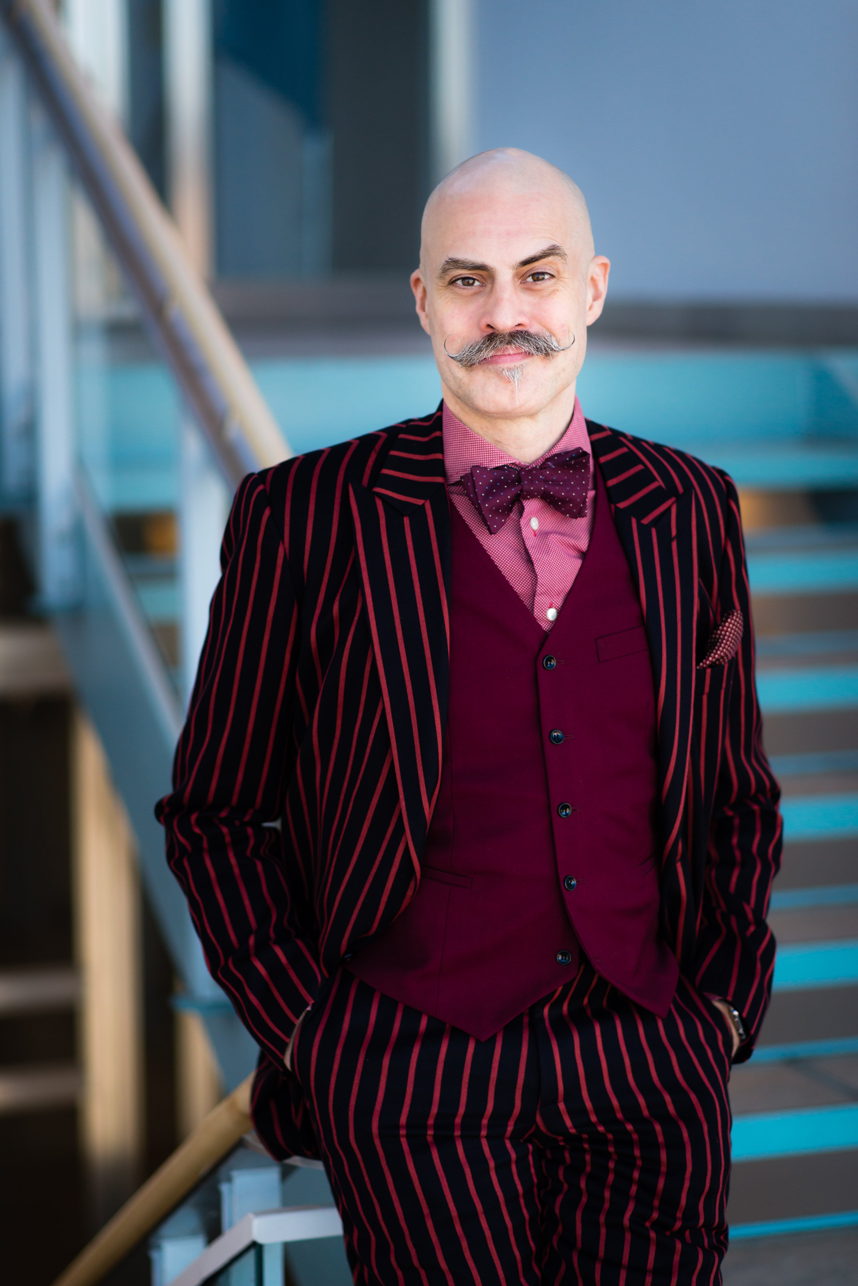 Porträtt på en man med mustasch klädd i en randig röd och svart kostym, vinröd väst, fluga och rosa skjorta.