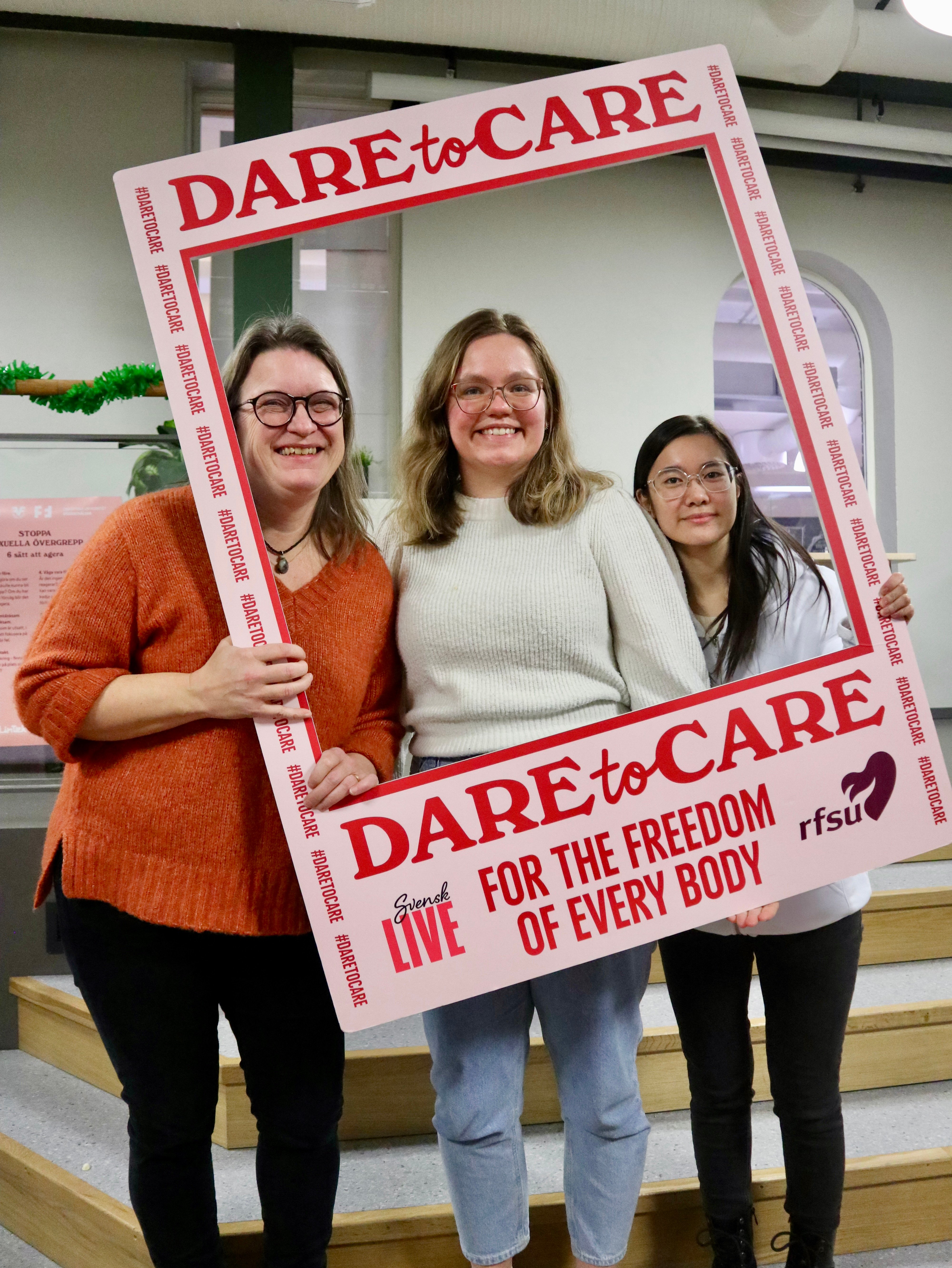 Tre leende personer poserar bakom en stor pappersram med texten Dare to Care.