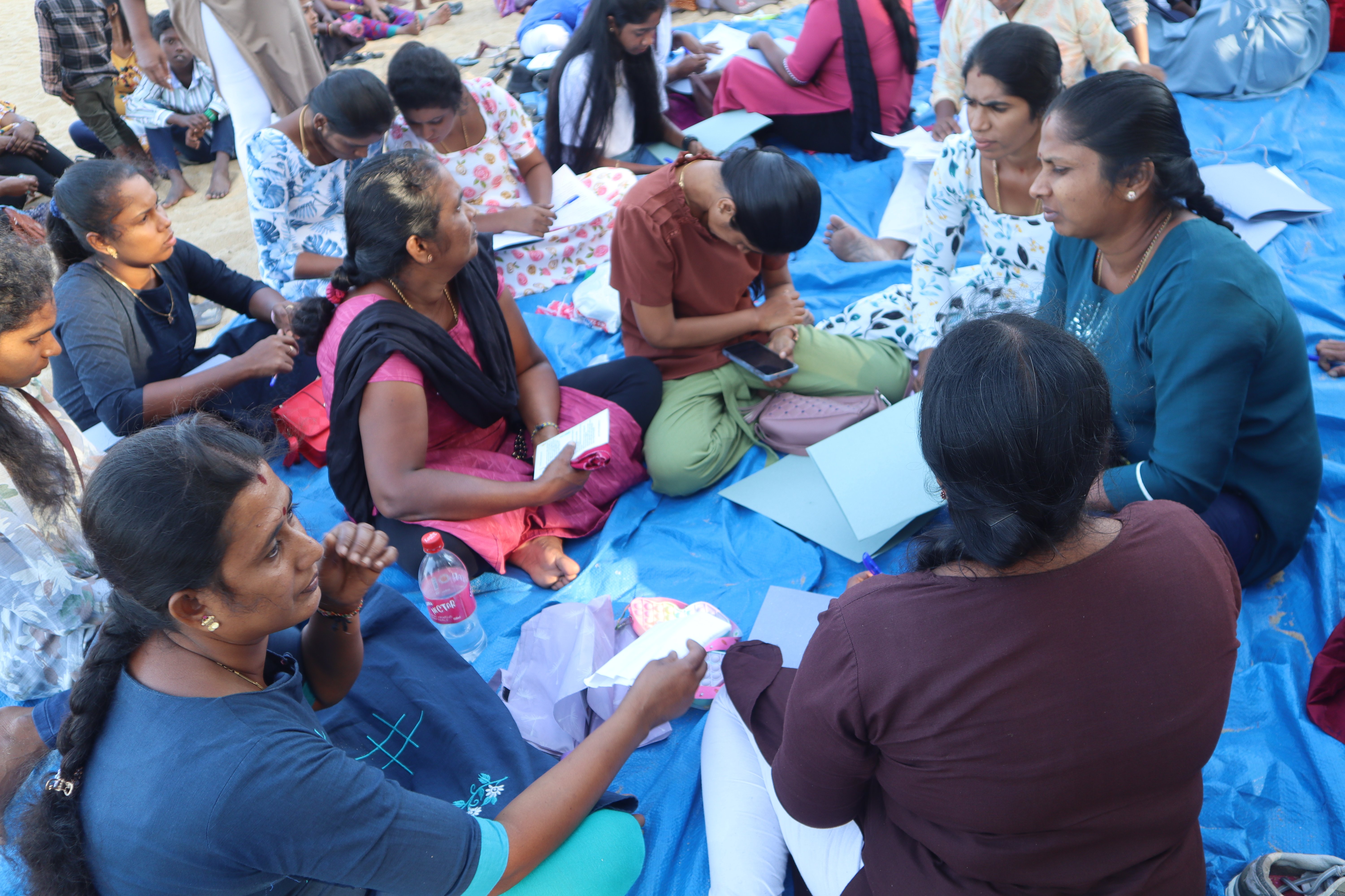 A group of women are seated closely in a circle on a blue tarp at the beach, engaged in a discussion while holding papers and notebooks.