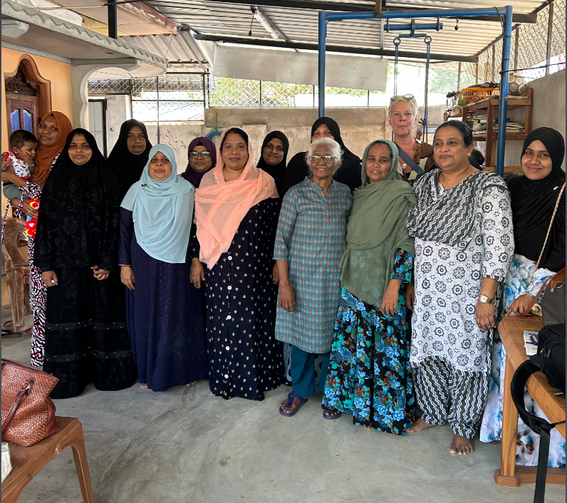 A group of women stand together for a photo under a covered outdoor patio. Many of the women are wearing colorful hijabs and patterned dresses, and one woman on the far left holds a small child.