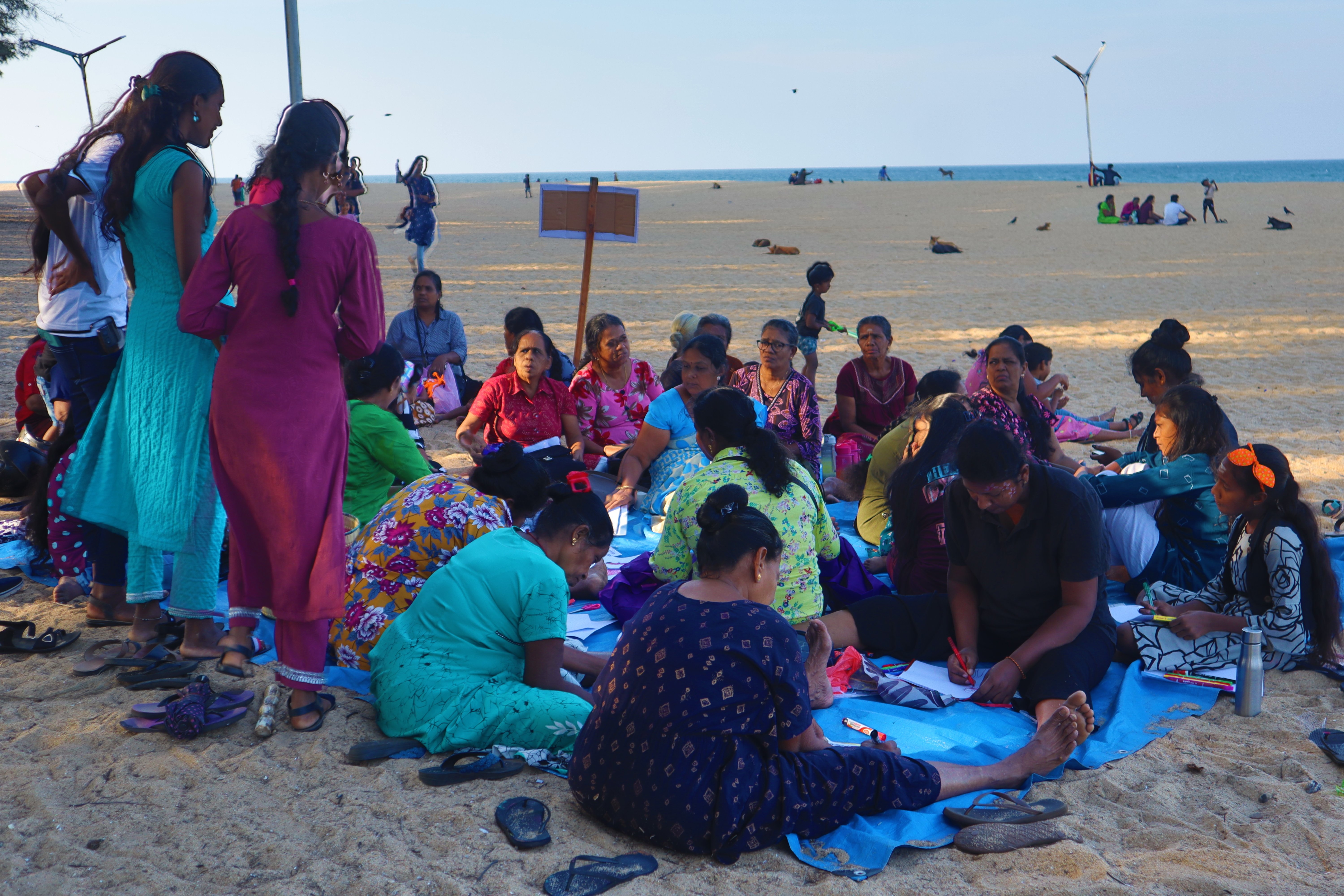 A large group of women are gathered on a blue tarp laid out on a sandy beach, with several individuals focused on writing or drawing on papers. The open ocean stretches across the background under a clear sky.