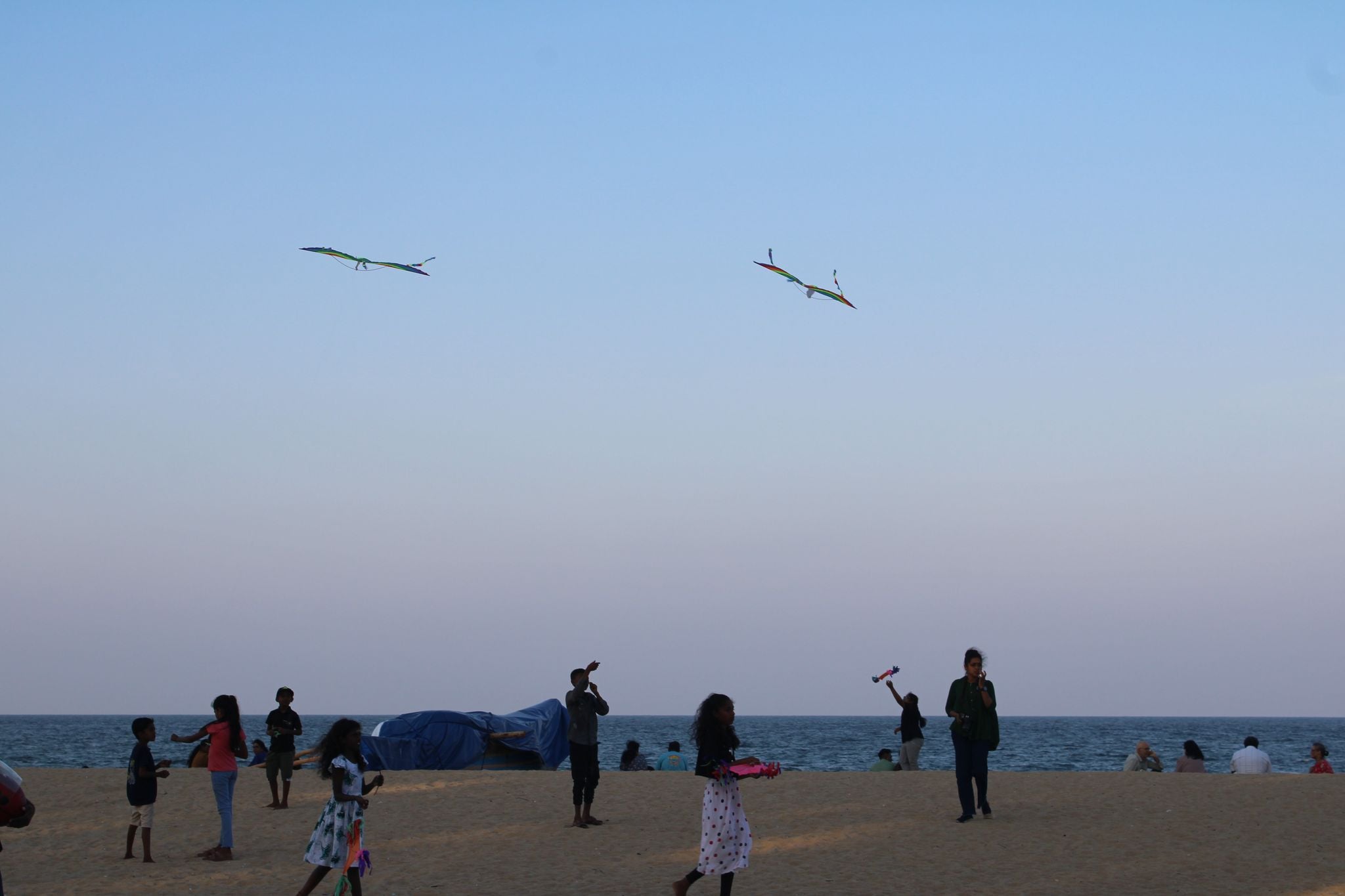 A few adults and children spread out across a sandy beach near the ocean under a pale evening sky. Two colorful kites fly high in the air above them.