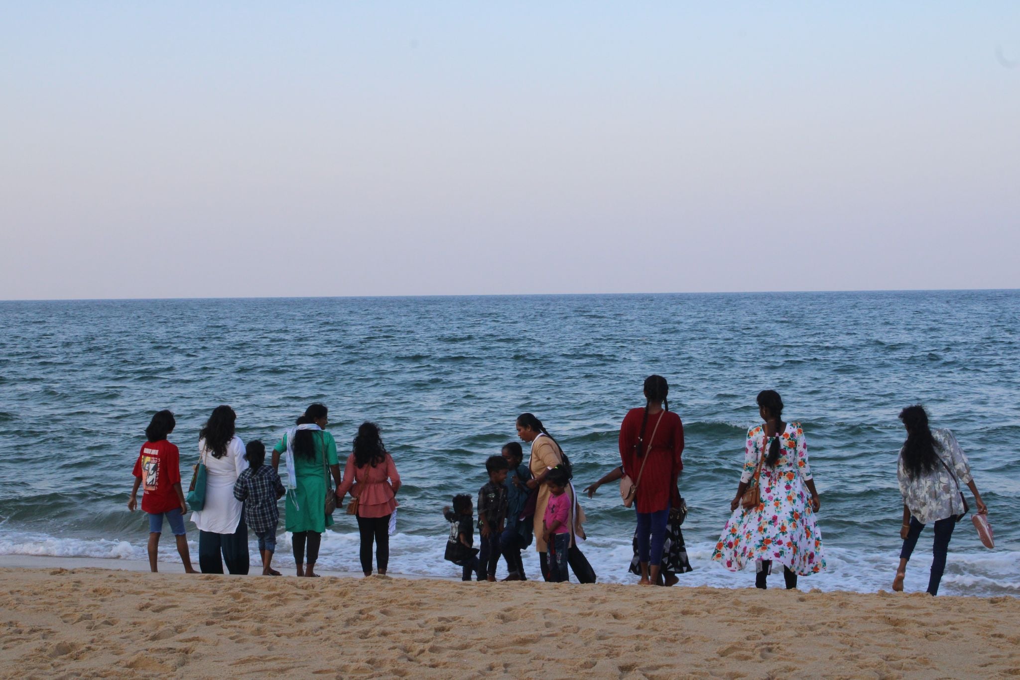 En grupp vuxna och barn står på en sandstrand och blickar ut över havet. De är vända bort från kameran och bär färgglada kläder, bland annat en blommig klänning, en grön tunika och en röd tröja. Himlen är blek och havet sträcker sig mot horisonten med små skummande vågor som rullar in mot stranden.