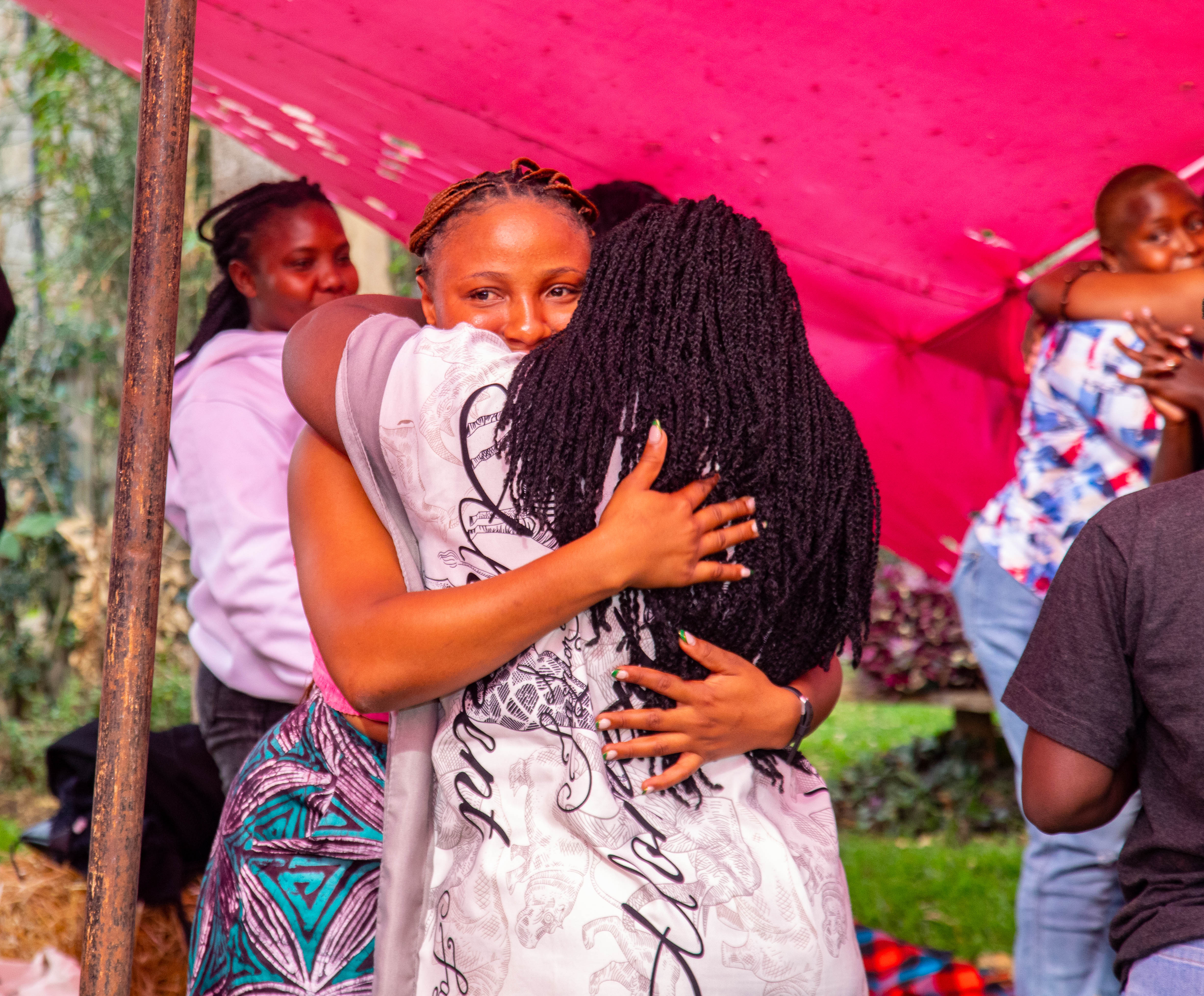 Two women share a warm, close embrace outdoors under a vibrant pink canopy. The woman facing the camera smiles gently with her arms wrapped tightly around her companion.