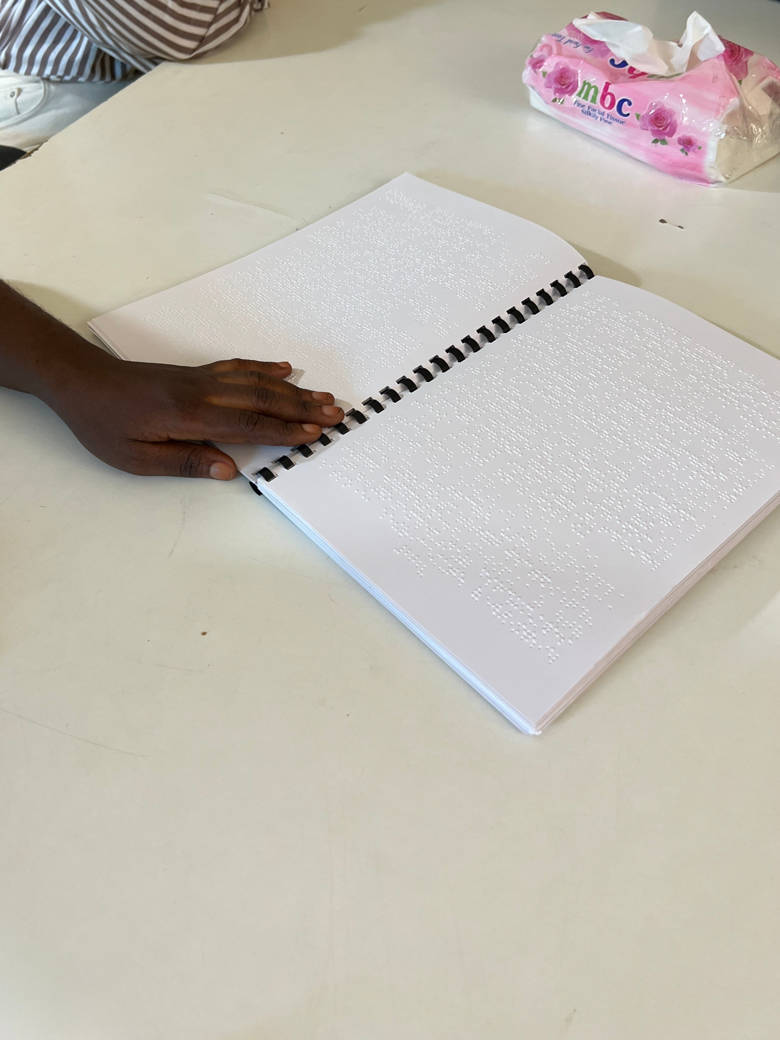 A close-up shows a person's hand resting on an open spiral-bound notebook filled with raised Braille text. A pink tissue box sits on the white table in the background.