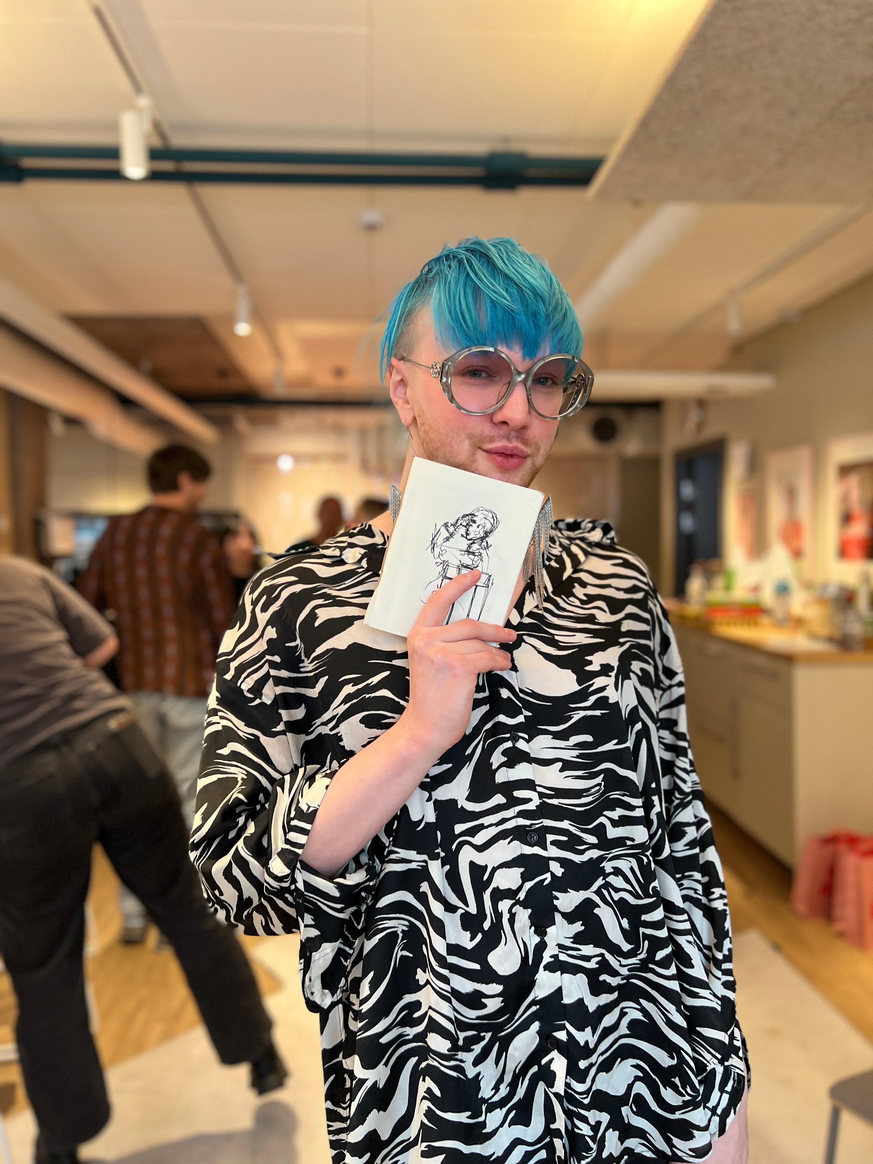 En person med blått hår och stora glasögon håller upp en liten teckning av en kramande figur.