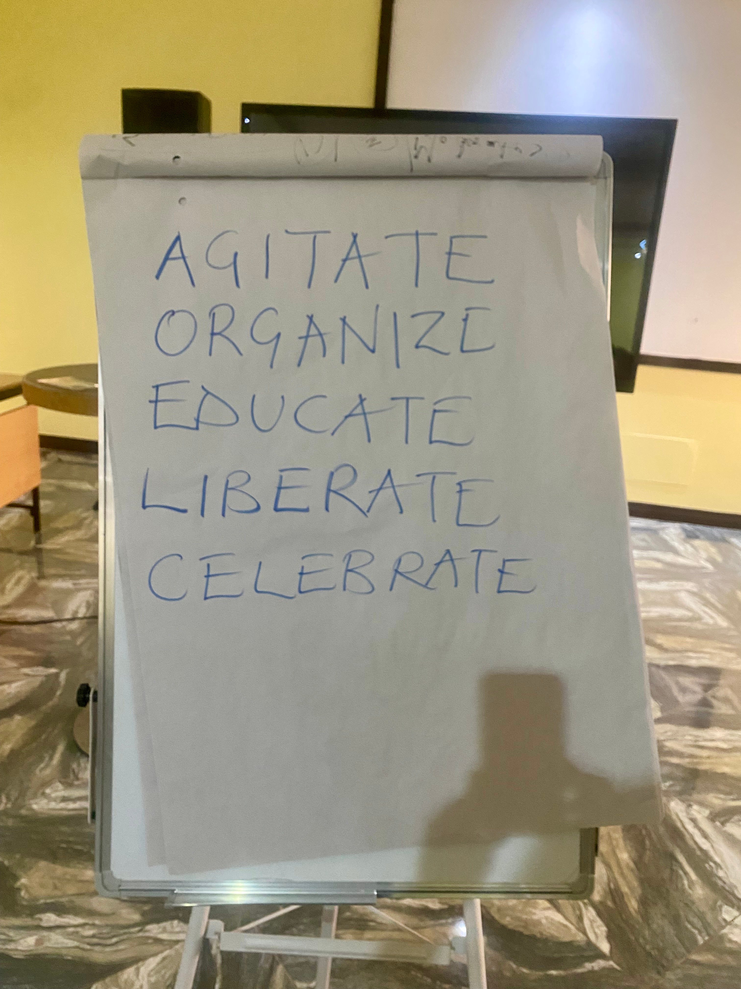 A white flipchart standing on an easel displays the words "AGITATE, ORGANIZE, EDUCATE, LIBERATE, CELEBRATE" handwritten in blue capital letters.